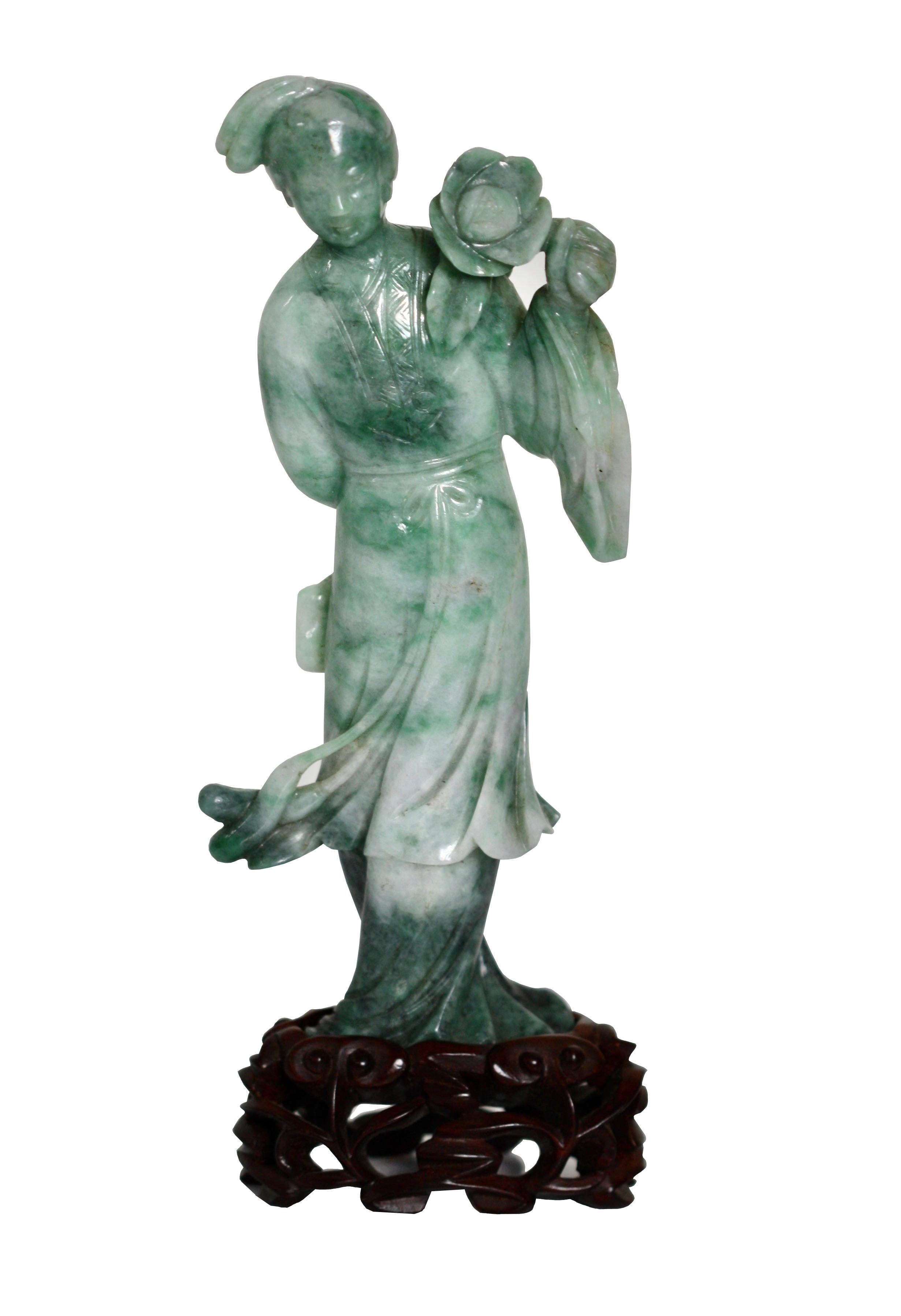 A Jadeite Figure of Guanyin, Chinese
Carved wearing long flowing robes, holding a flower, the lustrously polished stone with a long fissure ingeniously incorporated into the design, the back with some dark speckled inclusions, wood stand