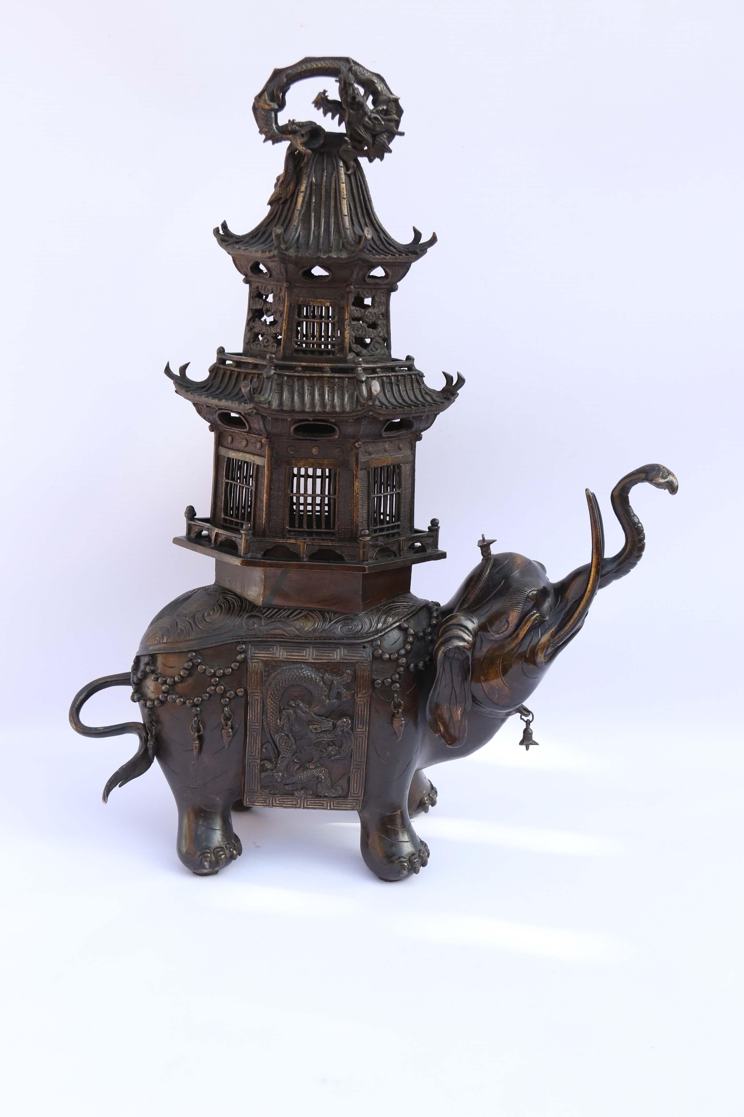 A highly decorative Japanese bronze study of ceremonial elephant wearing a elaborate saddlecloth adorned with jewels and a superbly detailed relief dragon decoration with a Greek key pattern surrounding it. The elephant is carrying a two storey