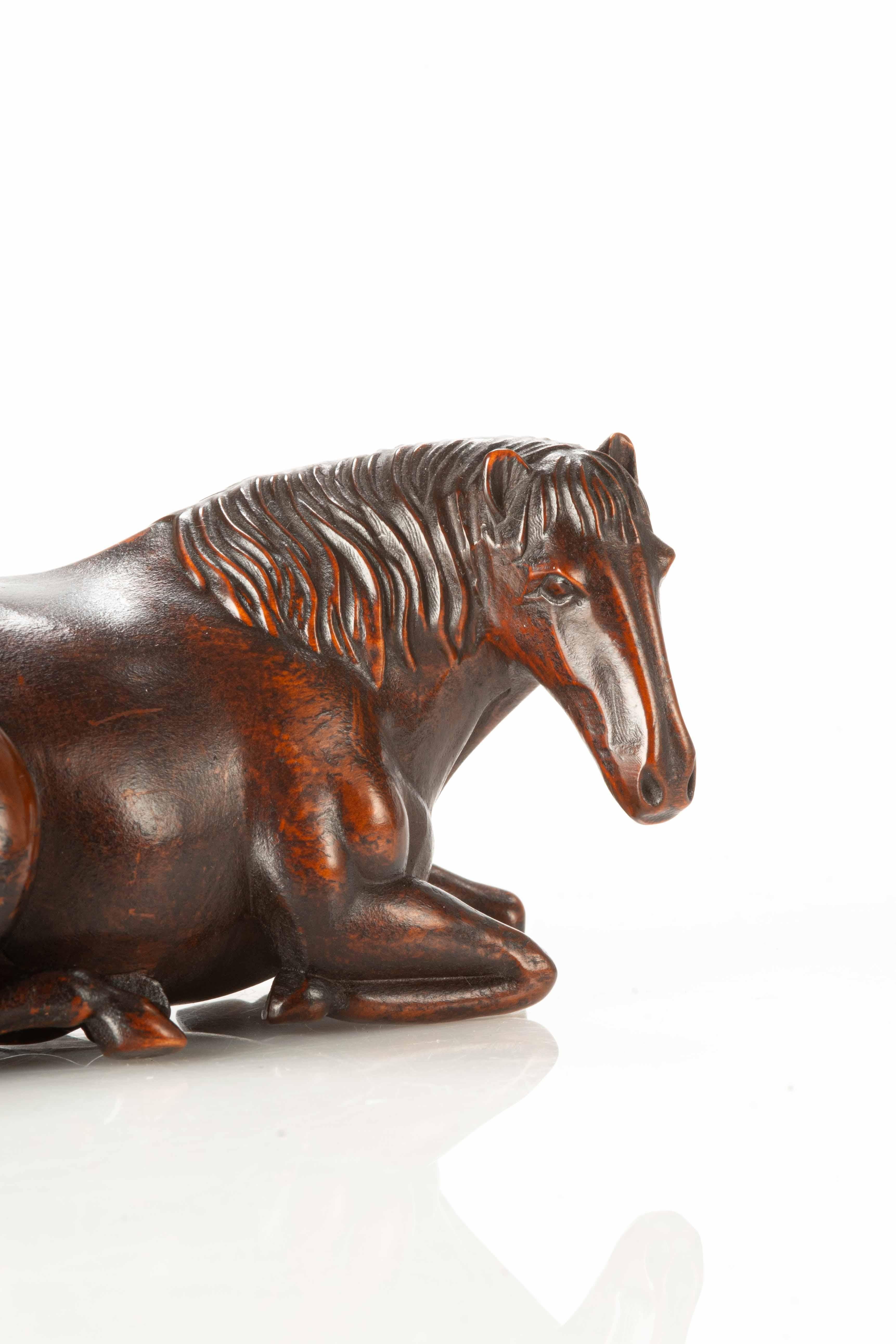 Okimono en bois représentant un cheval couché, finement sculpté dans les moindres détails. Les muscles, les sabots et la crinière sont rendus avec soin et art, capturant la vitalité du cheval dans une forme statique.

L'excellente patine qui