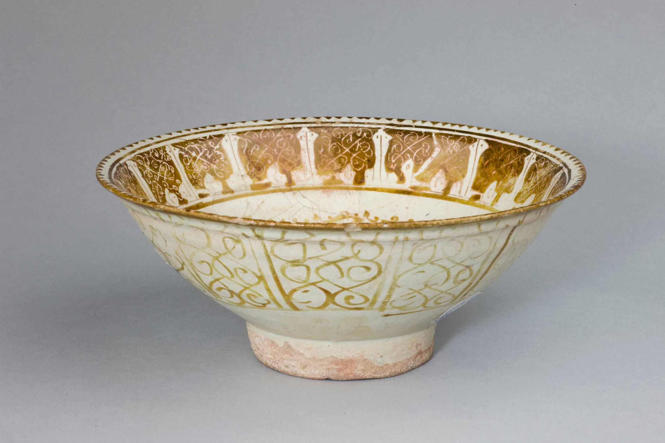 This intact lustre bowl is densely decorated in yellow lustre with a foliated motif in the centre, the well with dots and interlocking split palmettes radiating from a central medallion. A large band below the rim is heavily ornamented in drawn