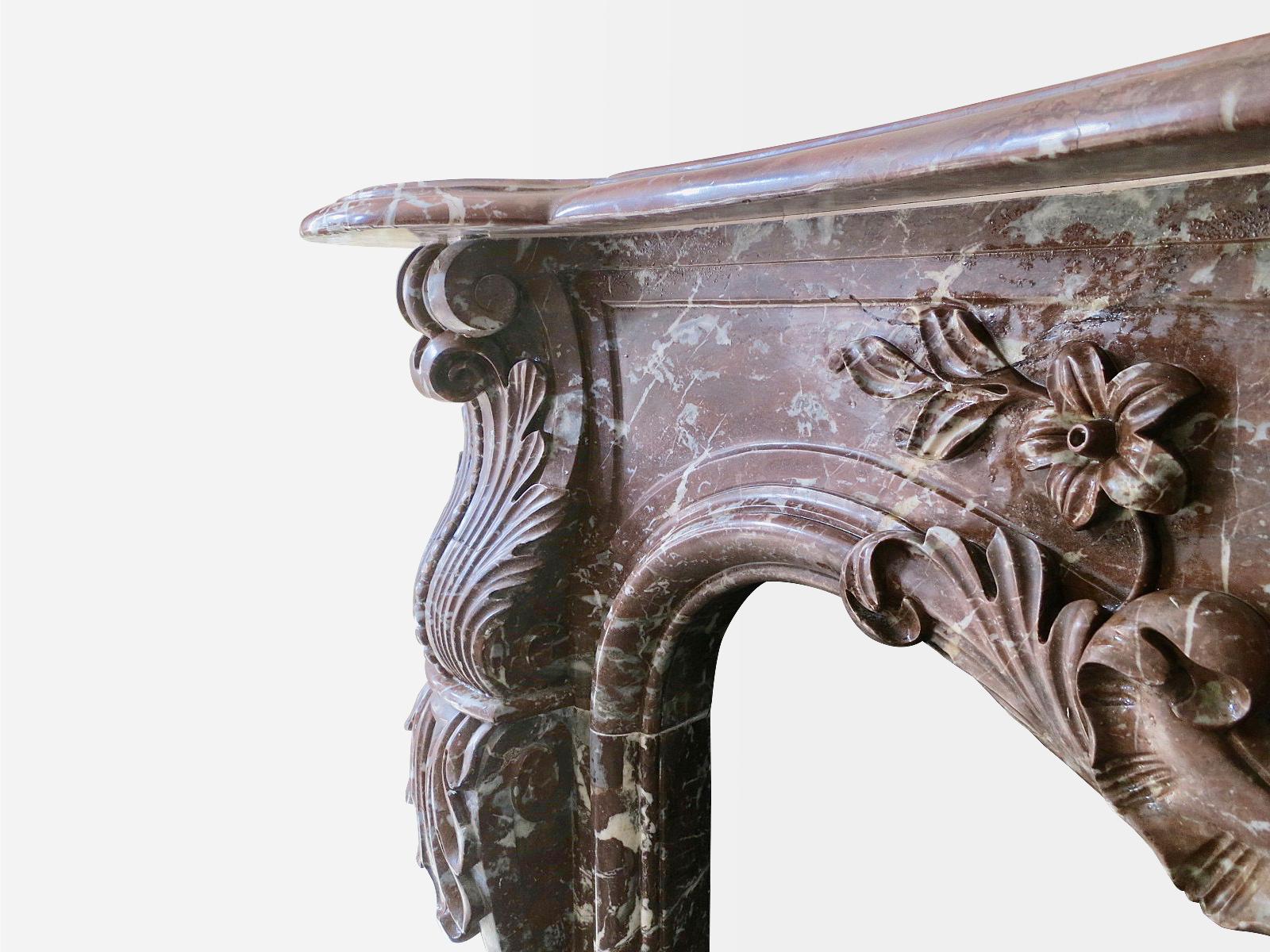 An ornately carved French antique fireplace in Rouge Royal marble. The jambs with scrolled Acanthus, surmounted by beaded C scrolled Acanthus end blocks. The centre cartouche of flowers and shell carved in high relief, with a large serpentine