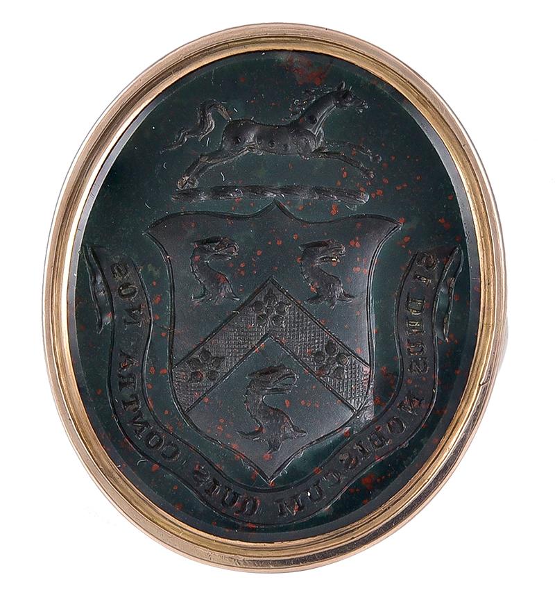 The very dark Bloodstone engraved with a Heraldic Coat of Arms comprising a shield within which are 3 Griffon Heads and 3 flowers in a hatched chevron. Above the shield is a galloping Horse on a ground line and below the shield within a scroll, is