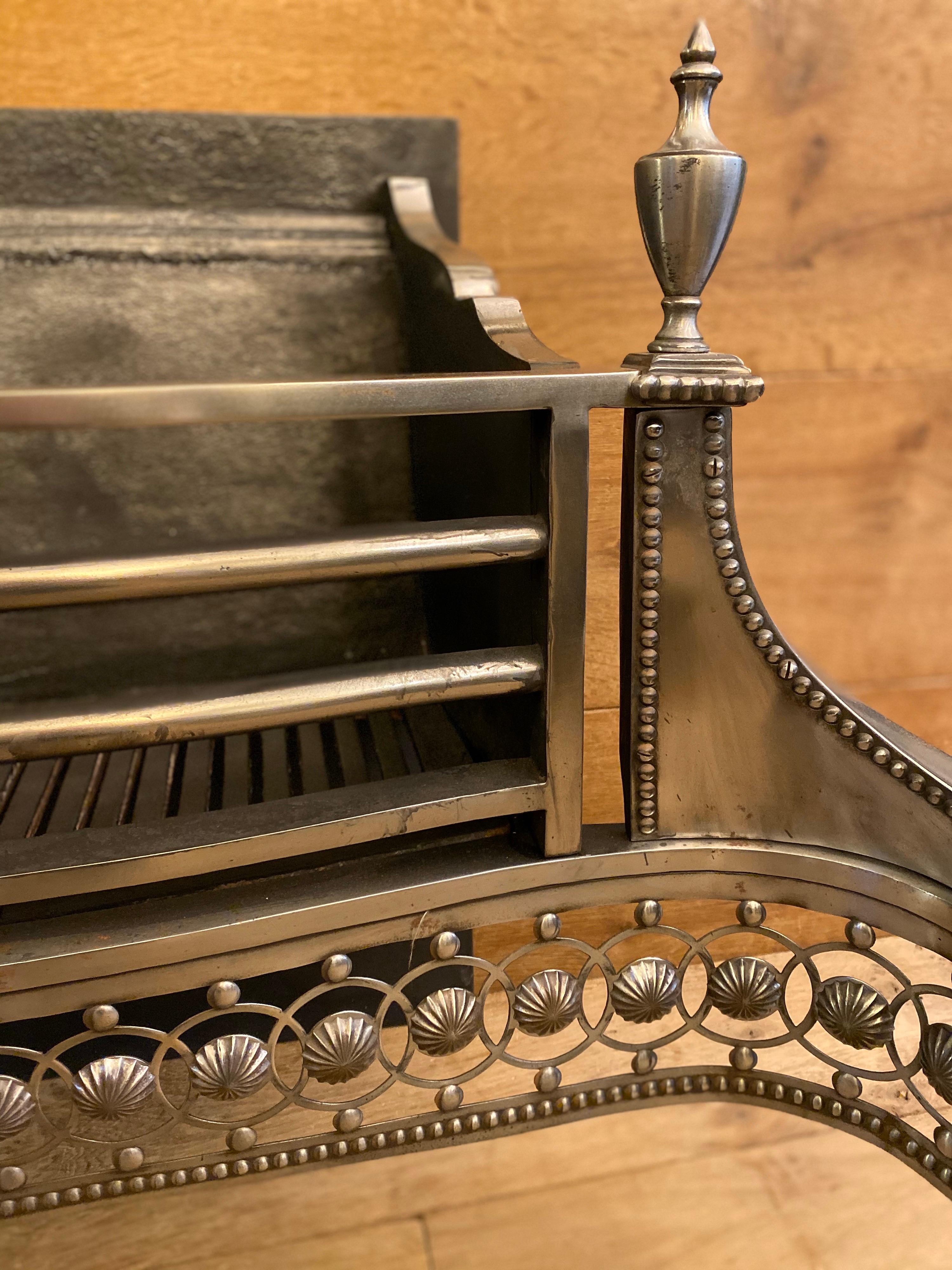 A large polished steel fire dog crate in the Georgian manner by the Carron foundry in Scotland. The tapered front legs surmounted by beaded edging and finial topped urns. Plain Beaded wings flank a bowed bar burning area, with a cut guilloche