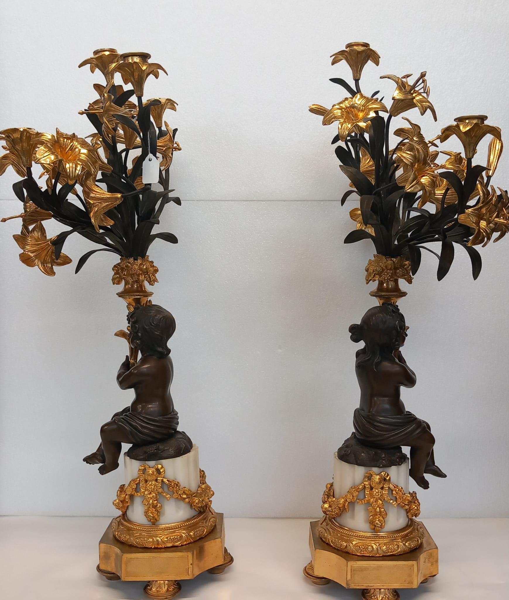 A large antique pair of French neoclassical candelabra in dark and gilt bronze depicting a young boy and a girl sitting on a white marble column, each holding a cornucopia of gilt tiger lilies with 5 candle holders on each. 
The gilt bronze bases