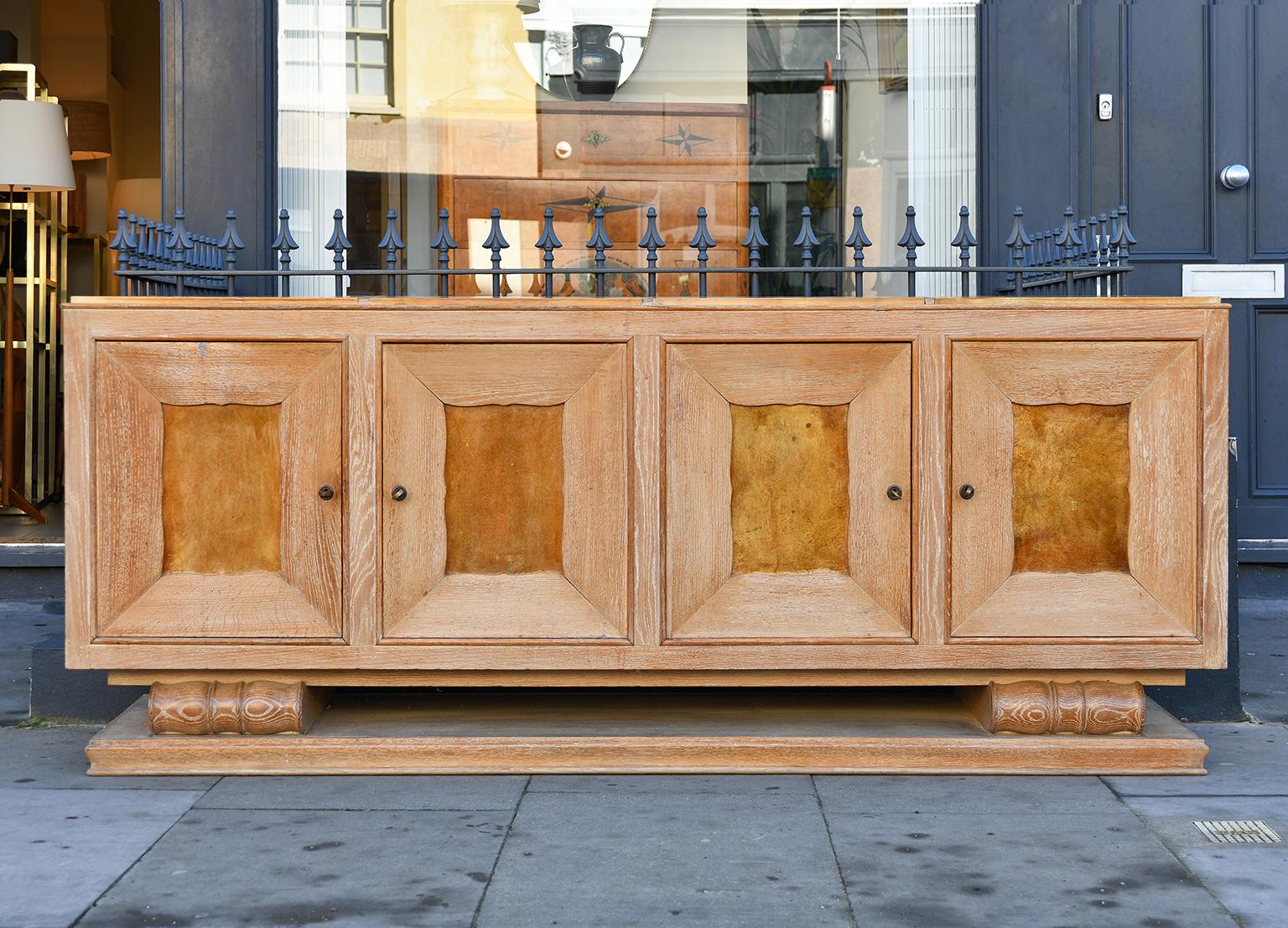 A large Art Deco limed oak and velum sideboard, by Perdoni Lombard (Décorateurs à Nice)
each of the four doors, adorned with velum, reveal an adjustable shelf and a drawer, the top is split into four sections of leather/velum also, resting on two