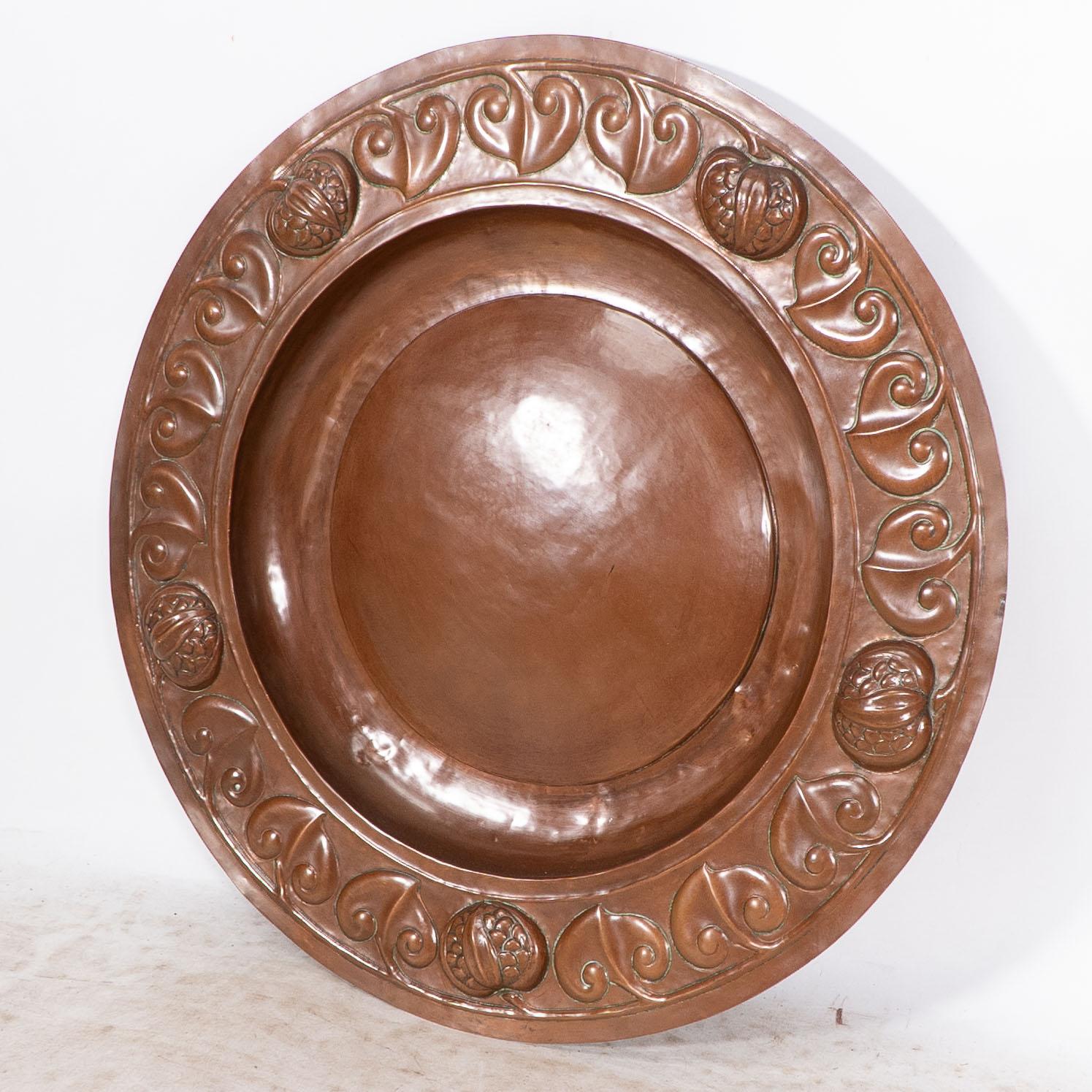 A large Arts and Crafts hand formed copper charger with stylized leaf and seed pod decoration to the border.