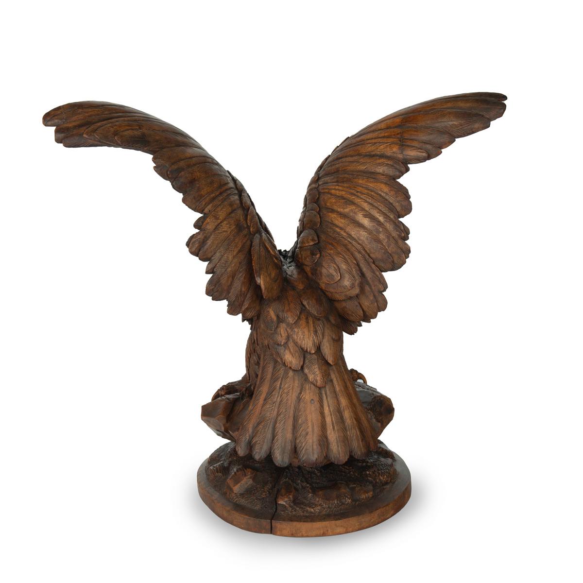 Grande sculpture en bois de noyer de la Forêt-Noire représentant un aigle, perché sur une chute de rochers, accroupi, les ailes déployées et la tête tournée vers la gauche, avec des détails tachés de noir sur les plumes et la tête.  yeux en verre