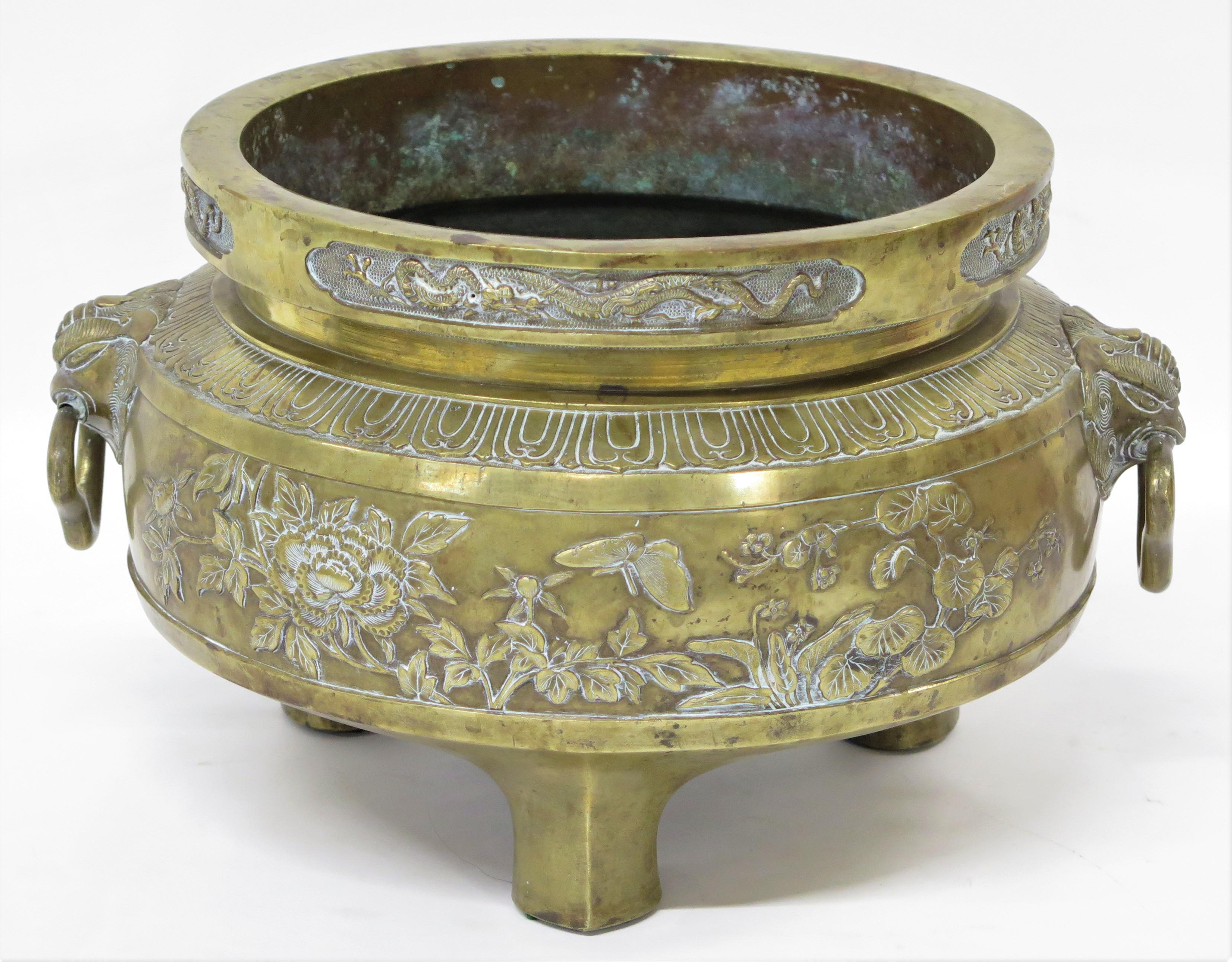 A large brass container three feet, dragons in relif insiad cartouches around top, floral relief band around center body. Rams heads with rings at sides. China. 19th Century.