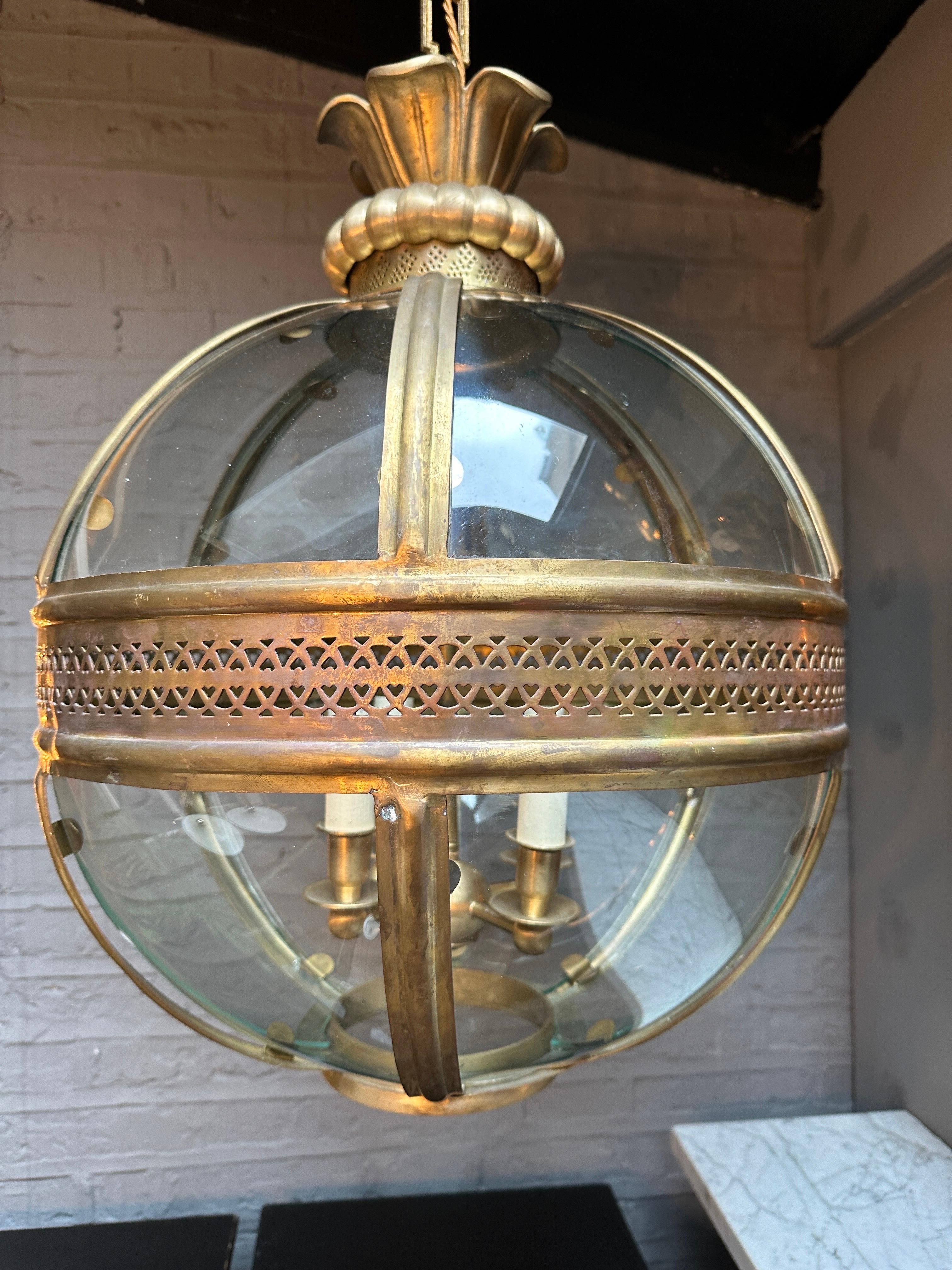 A large reclaimed hanging globe lantern in brass with distressed antique finish. The curved panels sitting with brass mouldings. The centre with pierced geometric pattern, four arm branch within. The canopy or rose of stiff leaf, gadroon and further