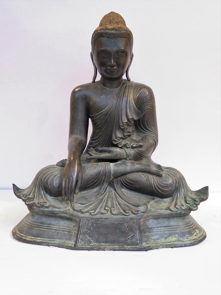 A bronze sculpture of a seated Buddha, Burmese.

 The Buddha sits upon a raised dais in virasana (half lotus position) gazing straight ahead, one hand in his lap, the other hand draped gracefully over his knee, reaching down, but not yet touching