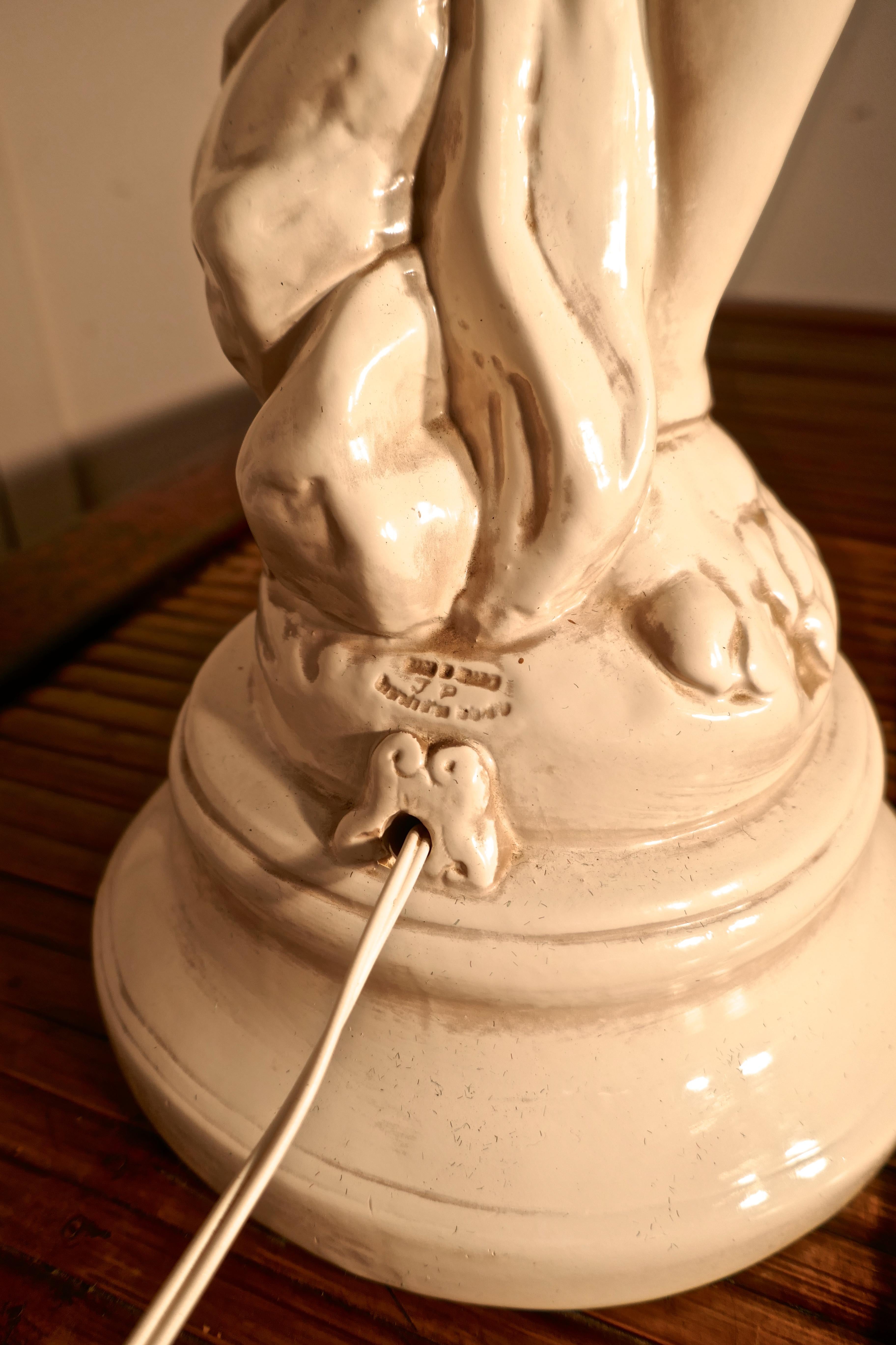 A large ceramic table lamp in the form of a Cherub or Putti

This charming figure is in an ivory colour and is set on a circular plinth, the cherub is holding a glass Art Deco flame lamp shade set in brass collar
The lamp is in very good