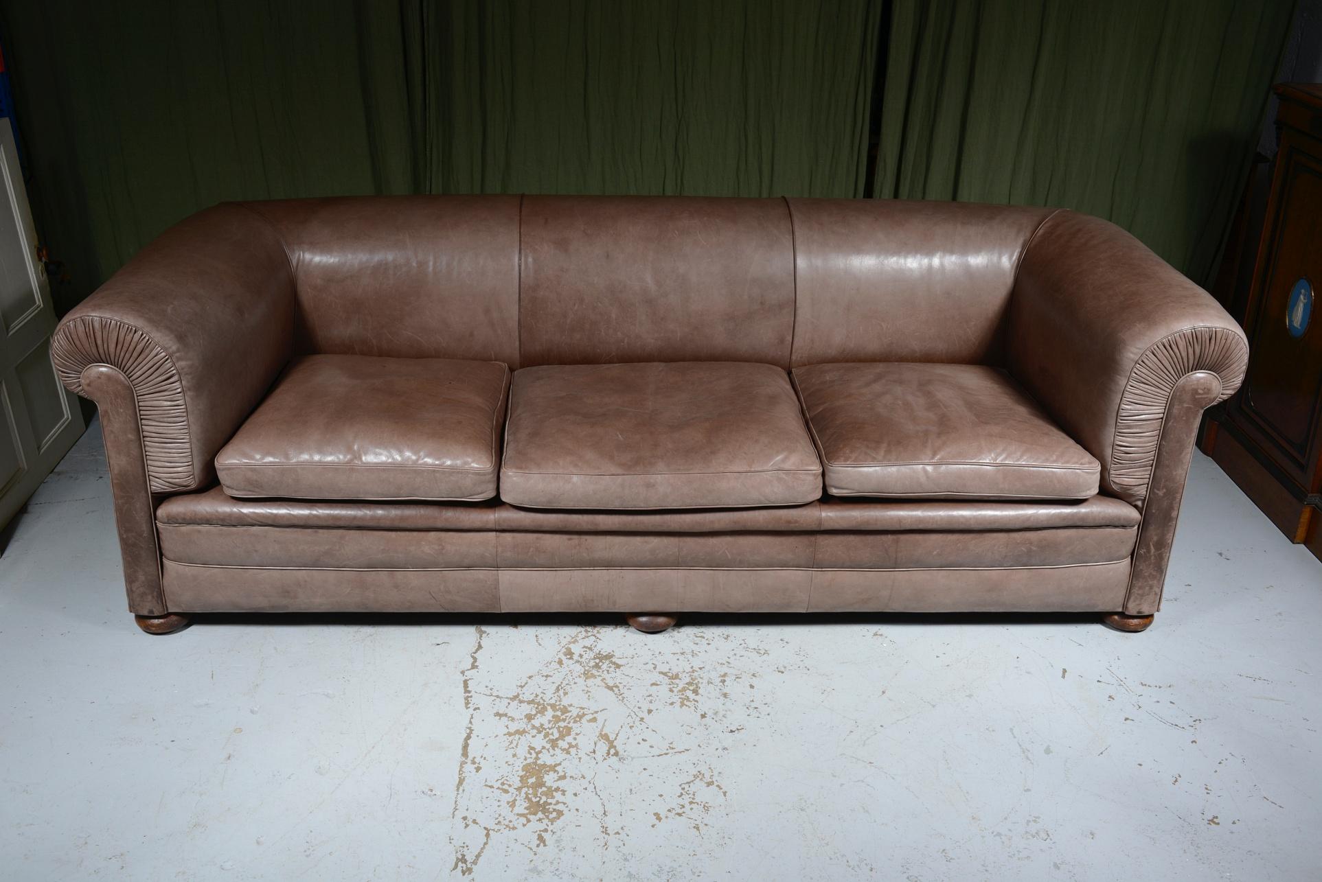 A very comfortable eight foot Edwardian chesterfield sofa, upholstered in supple light brown leather which has faded to a pleasant grey colour, with down filled cushions, on three oak bun feet to the front with sunken brass castors.