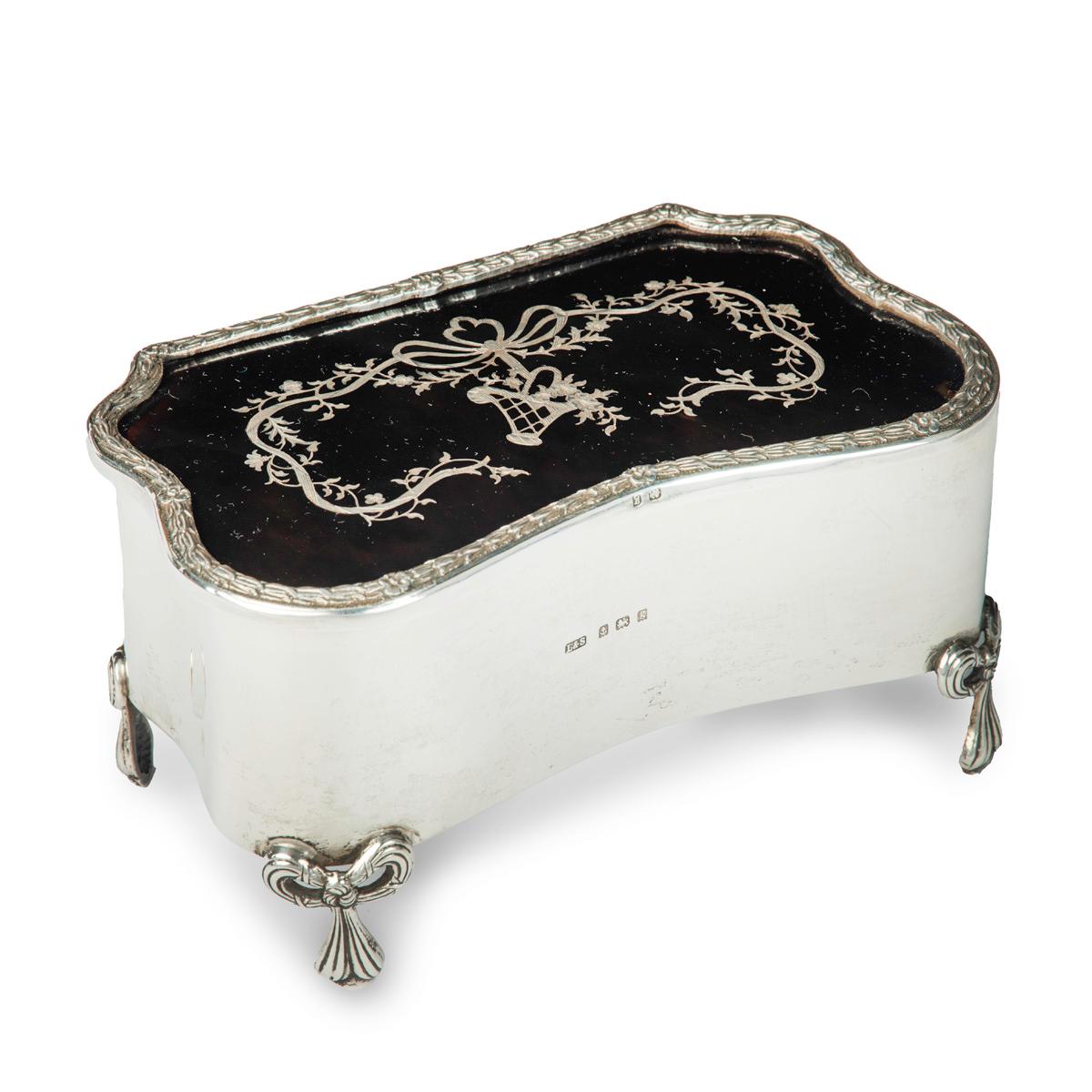 A large Edwardian shaped rectangular silver and tortoiseshell piqué box, the hinged lid with a dainty basket suspended from a bow with long ribbons entwined with flowering tendrils, all within a laurel and flowerhead border, raised on four tied-bow