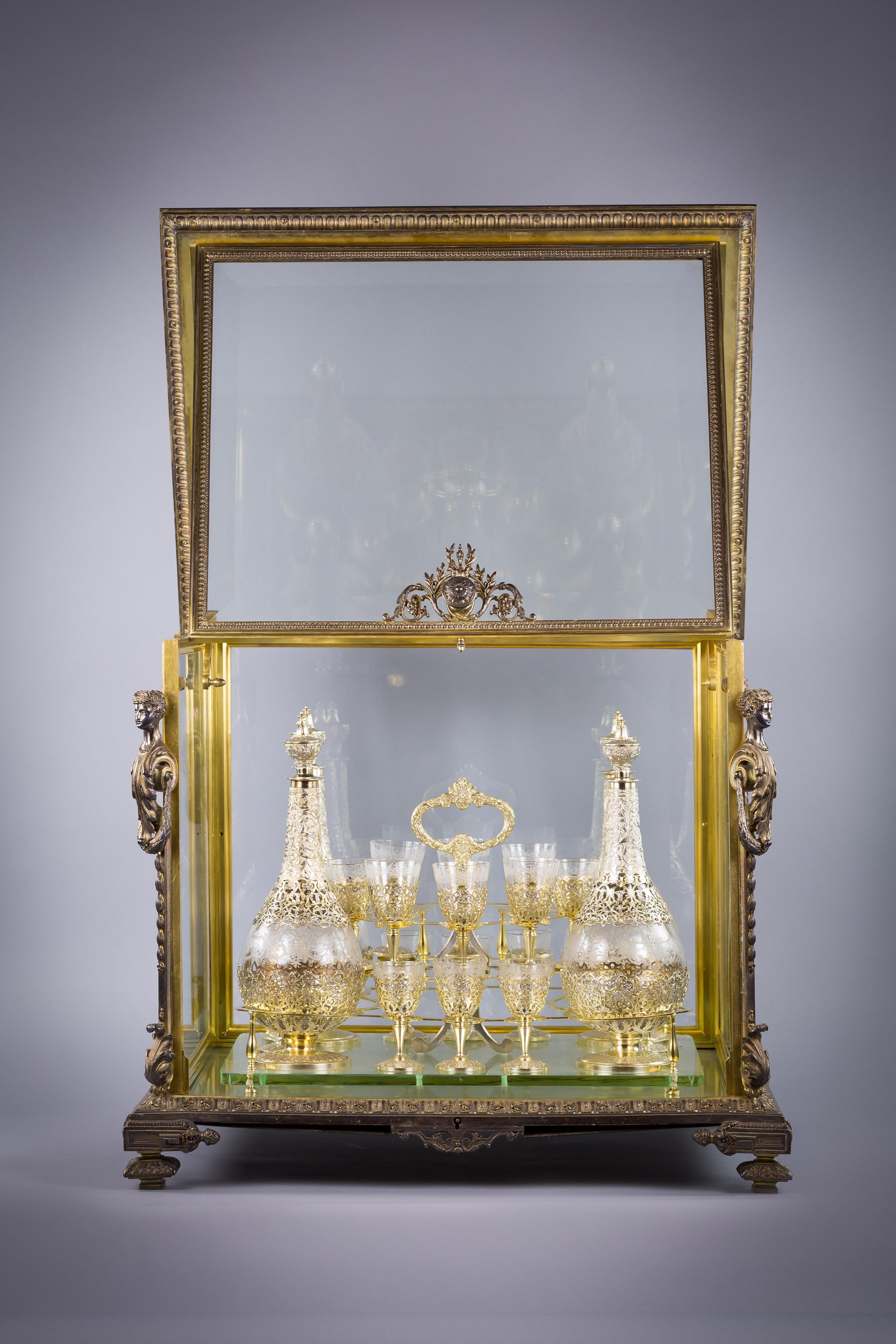 The rectangular case with bevelled glass sides and top, flanked to each angle by a female caryatid. the mirror interior fitted with a gilt silver mounted removable glass tray holding four silver gilt and engraved glass decanters and stoppers and