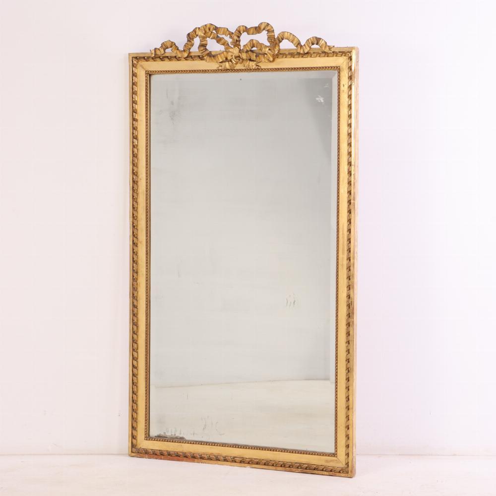 Featuring a delicately carved ribbon crest at the top, this substantial rectangular frame retains its warm gilded finish. Typical of late 19th-century French decorative arts, the mirror blends Rococo Revival elegance with restrained