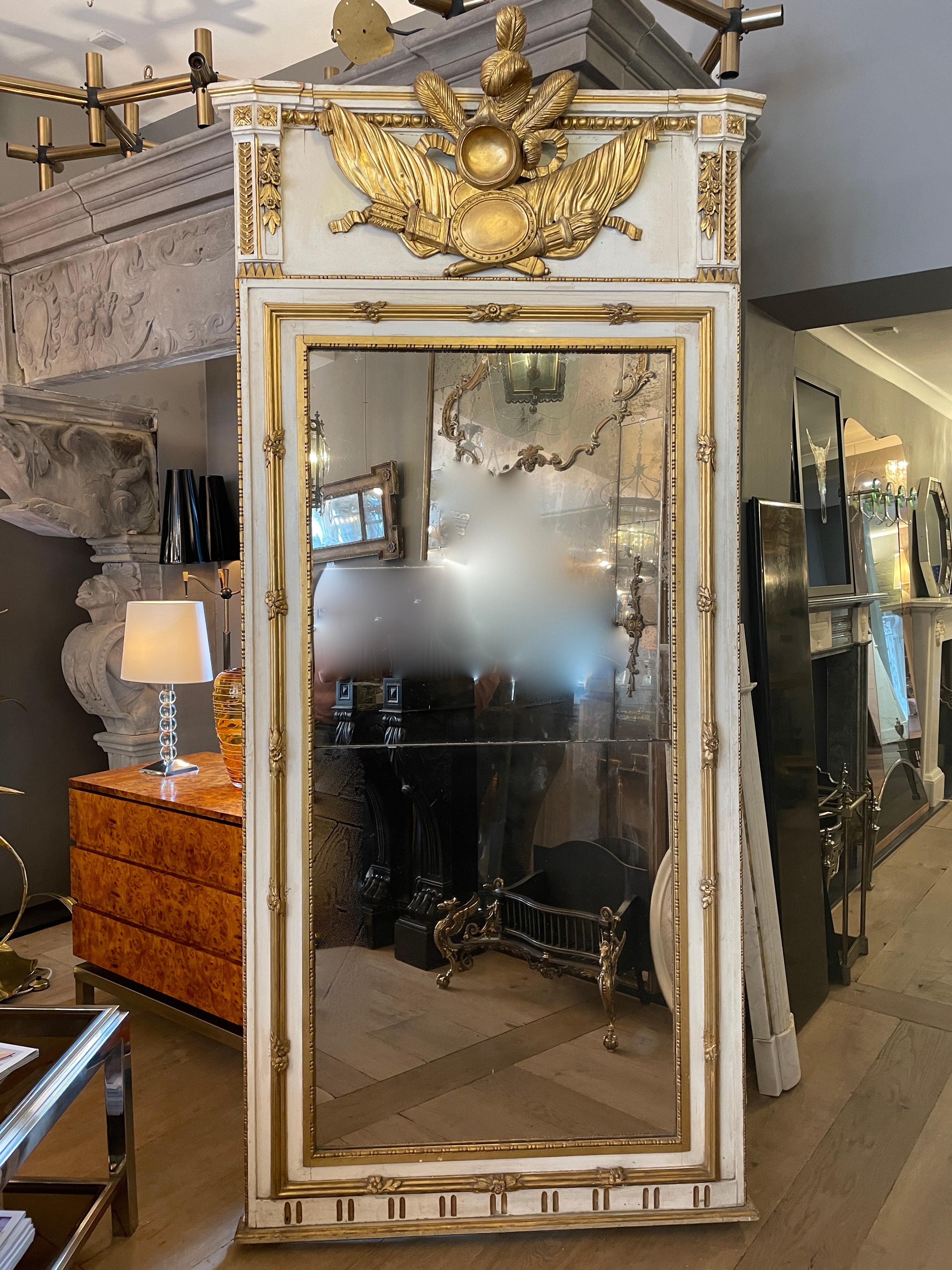 A 19th century antique Trumeau mirror in parcel gilt finish with original split mirror plate and carved wood pediment. Carved and mounted the cartouche of feathers, drapery, medallion, torch and quiver. The top decorated with rosettes, descending