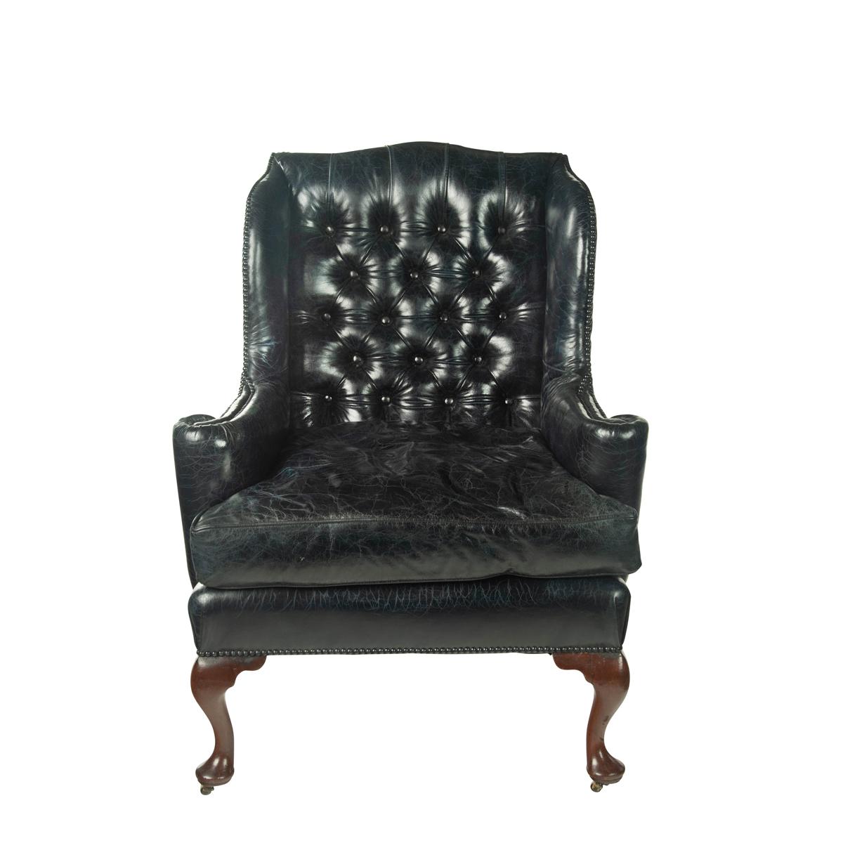 Un grand fauteuil à oreilles de George III, avec un dossier en forme de bouton profond et des accoudoirs en volute, tapissé à nouveau de cuir bleu-noir usé, reposant sur quatre pieds cabriole avec les roulettes d'origine.  Anglais, vers