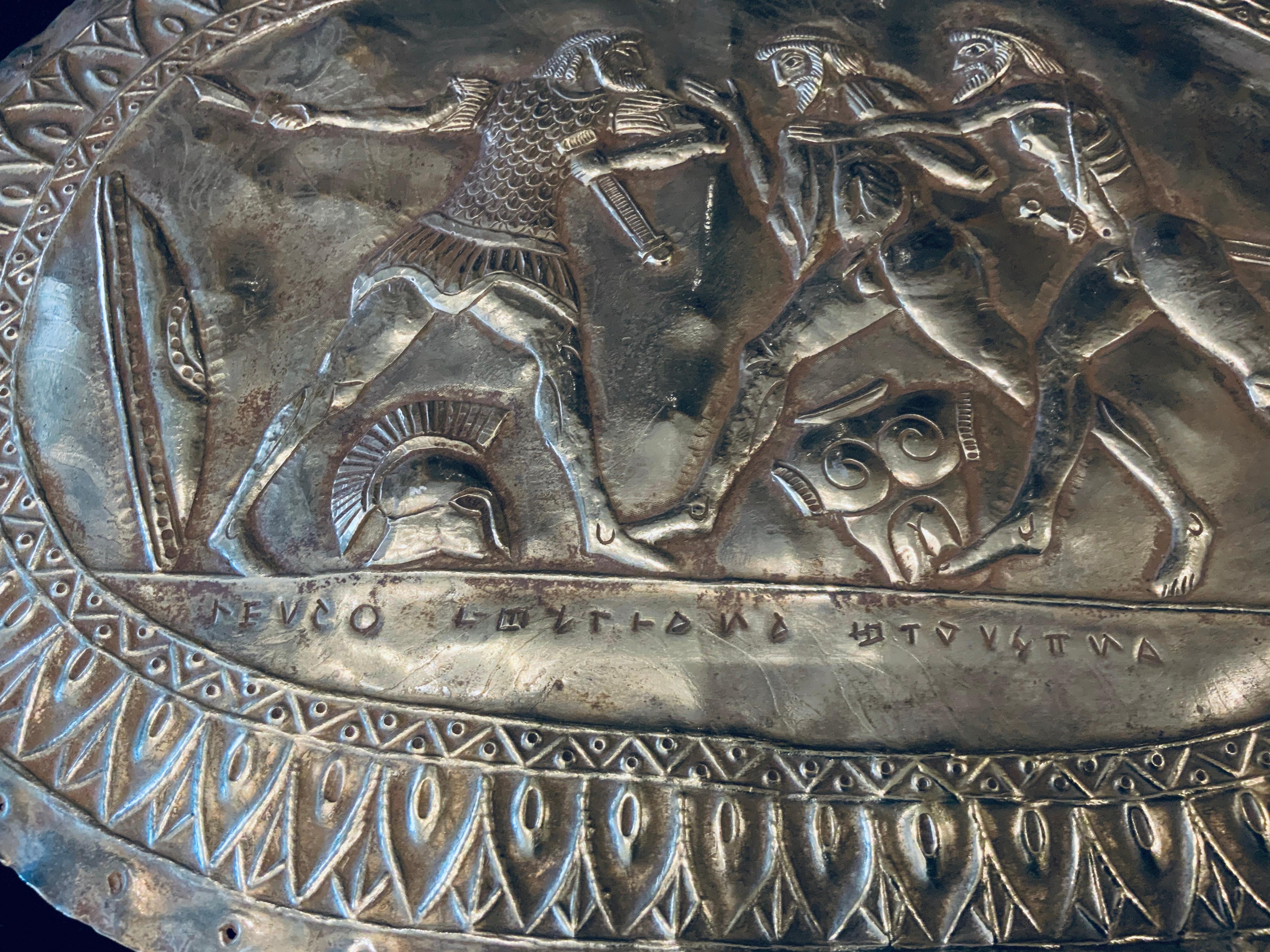 This is a large Greek style repousse oval metal plaque depicting a scene of a Spartan warrior trying to defend himself against two nudes warriors from another tribe. There are two shields standing at each side of them. There are also a Spartan