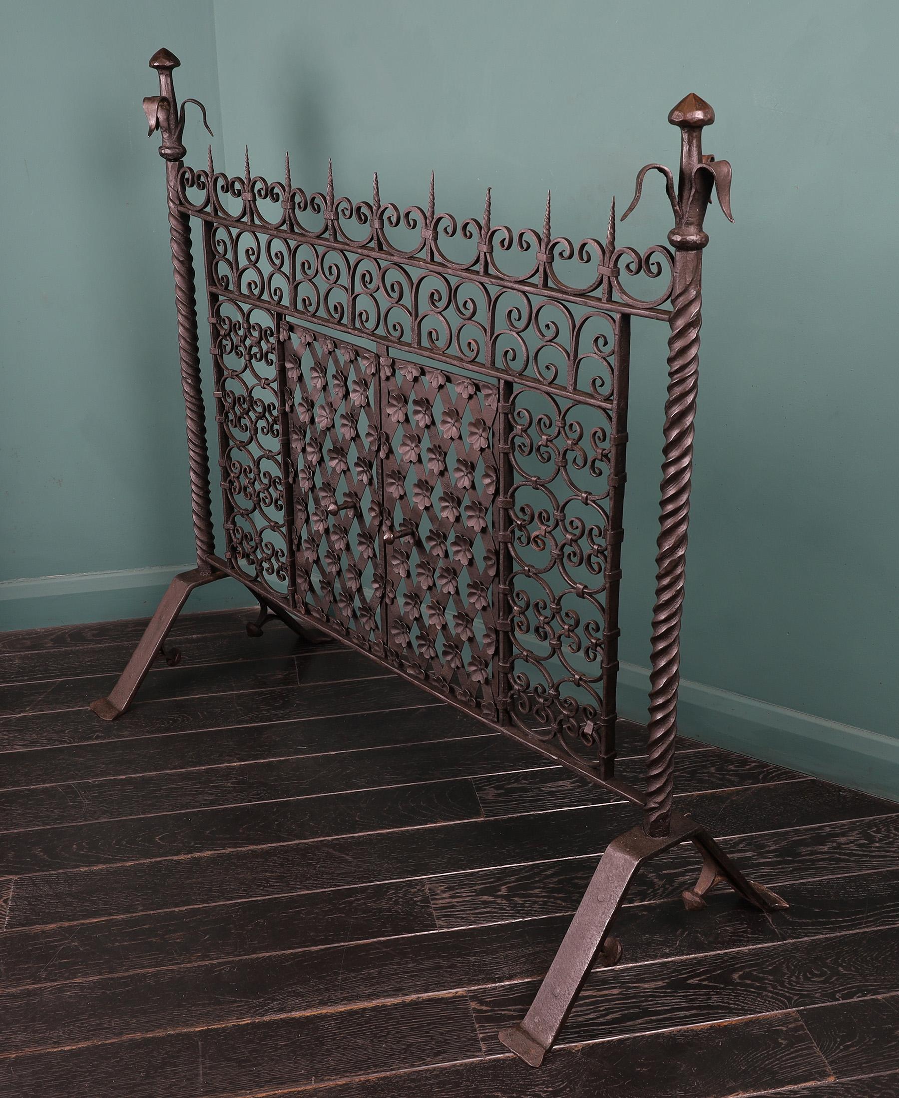 A Large Hand-Wrought Fire Screen with central access doors. The stylised flower finial above heavy uprights with twist detail standing on arched feet. The fire panel is made up of a pair of lattice and flower doors surrounded by linked c-scrolls. A