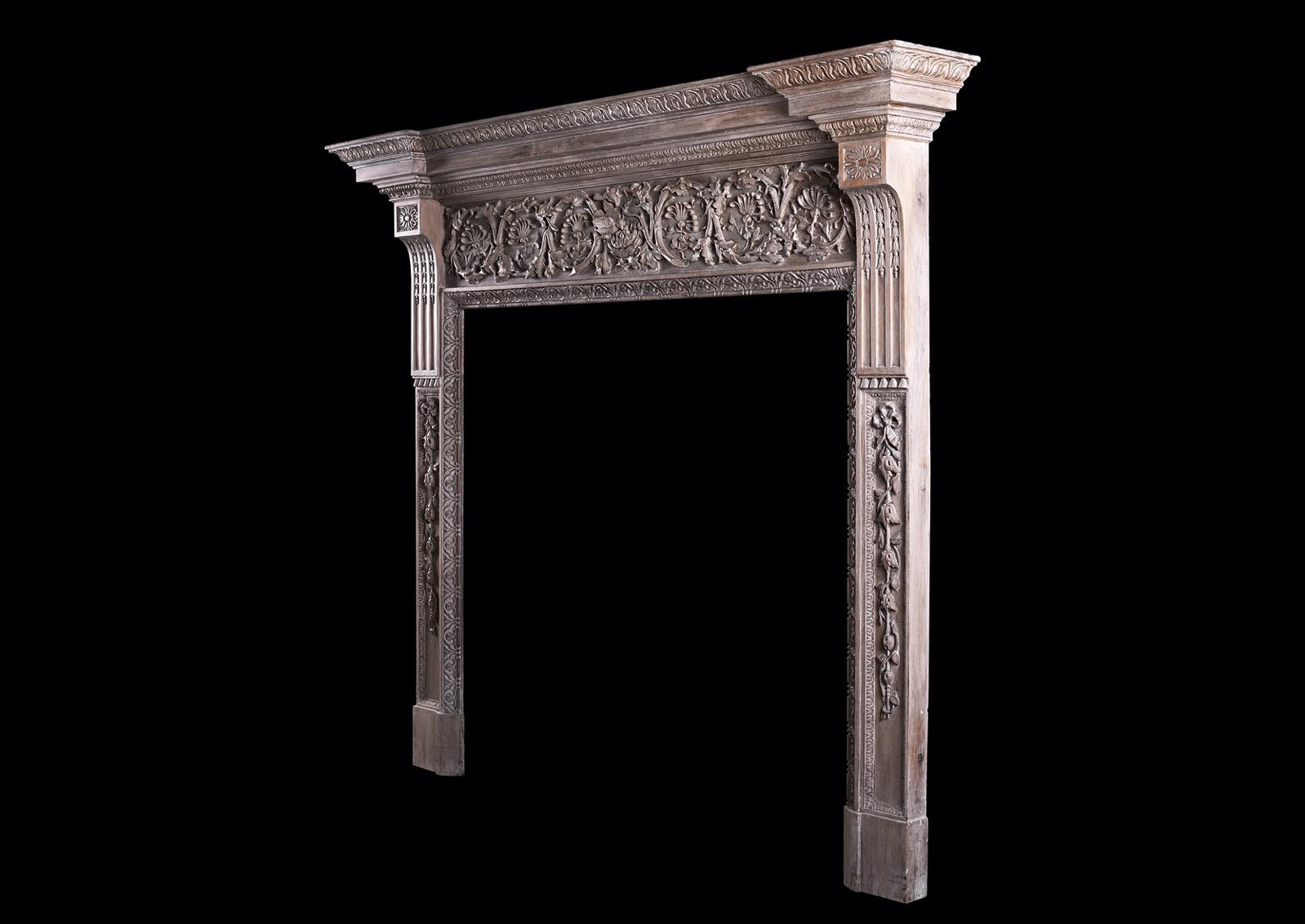 A large and impressive wood fireplace. The panelled jambs with carved ribbon and bellflowers, surmounted by brackets inlaid with husks with oval paterae above. The frieze with carved leaf work and foliage throughout with birds to centre. The