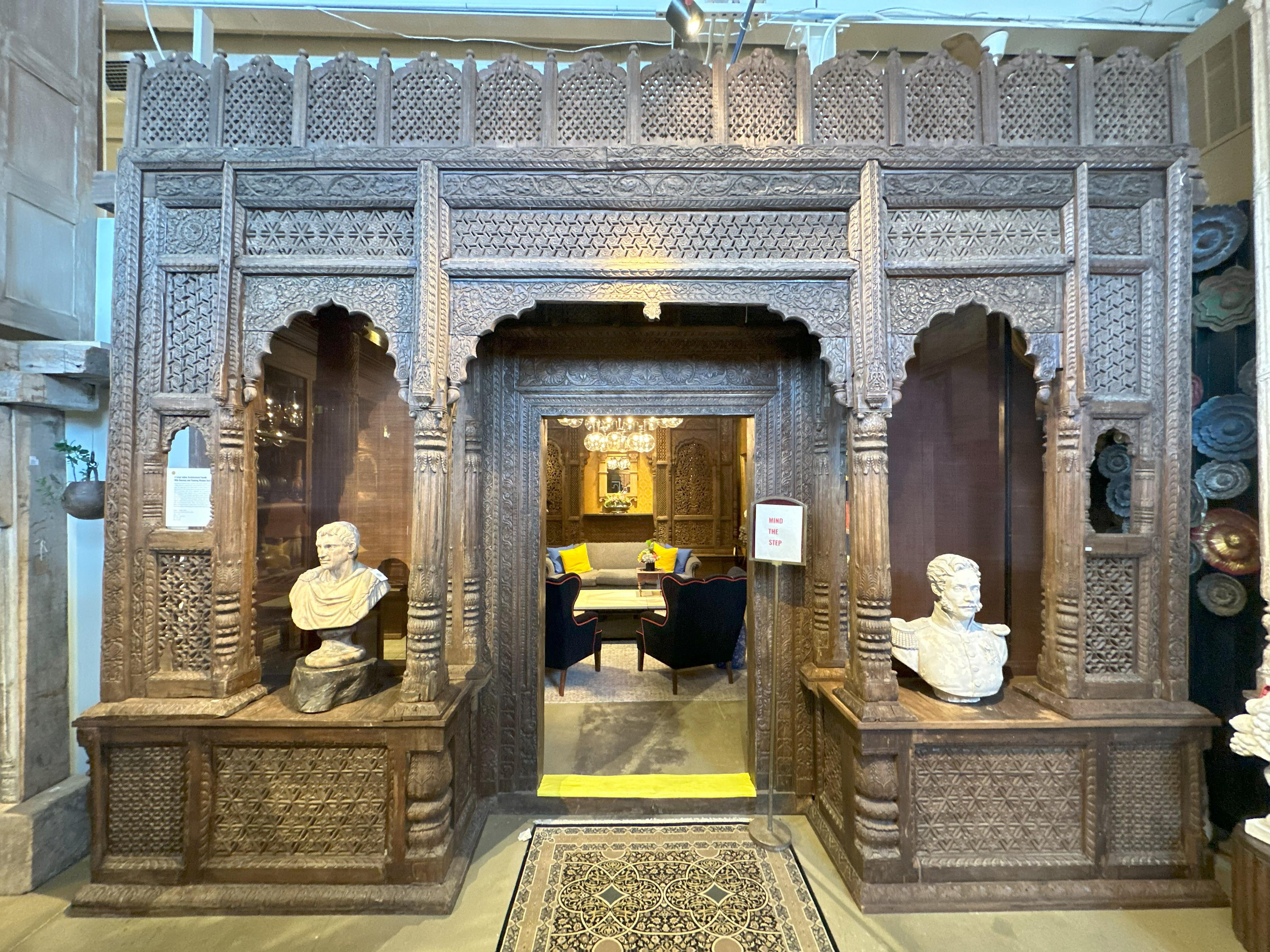 This stunning Indo-Saracenic wooden entrance gate, salvaged from a c.1920s mansion in Gujarat, India, features flanking built-in window seats. The upper part of the facade boasts a series of chhatri dome panels adorned with Champa flower and peacock