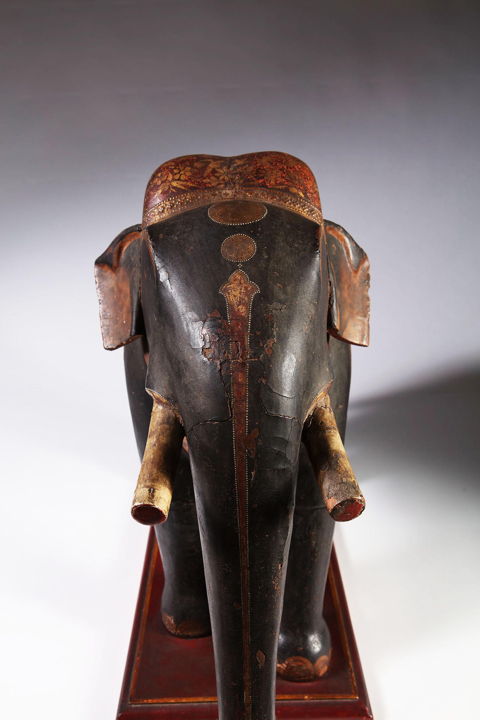 A large polychrome carved wooden processional elephant decorated with foliate head dress and foliate designs throughout, standing on a rectangular wooden plinth.