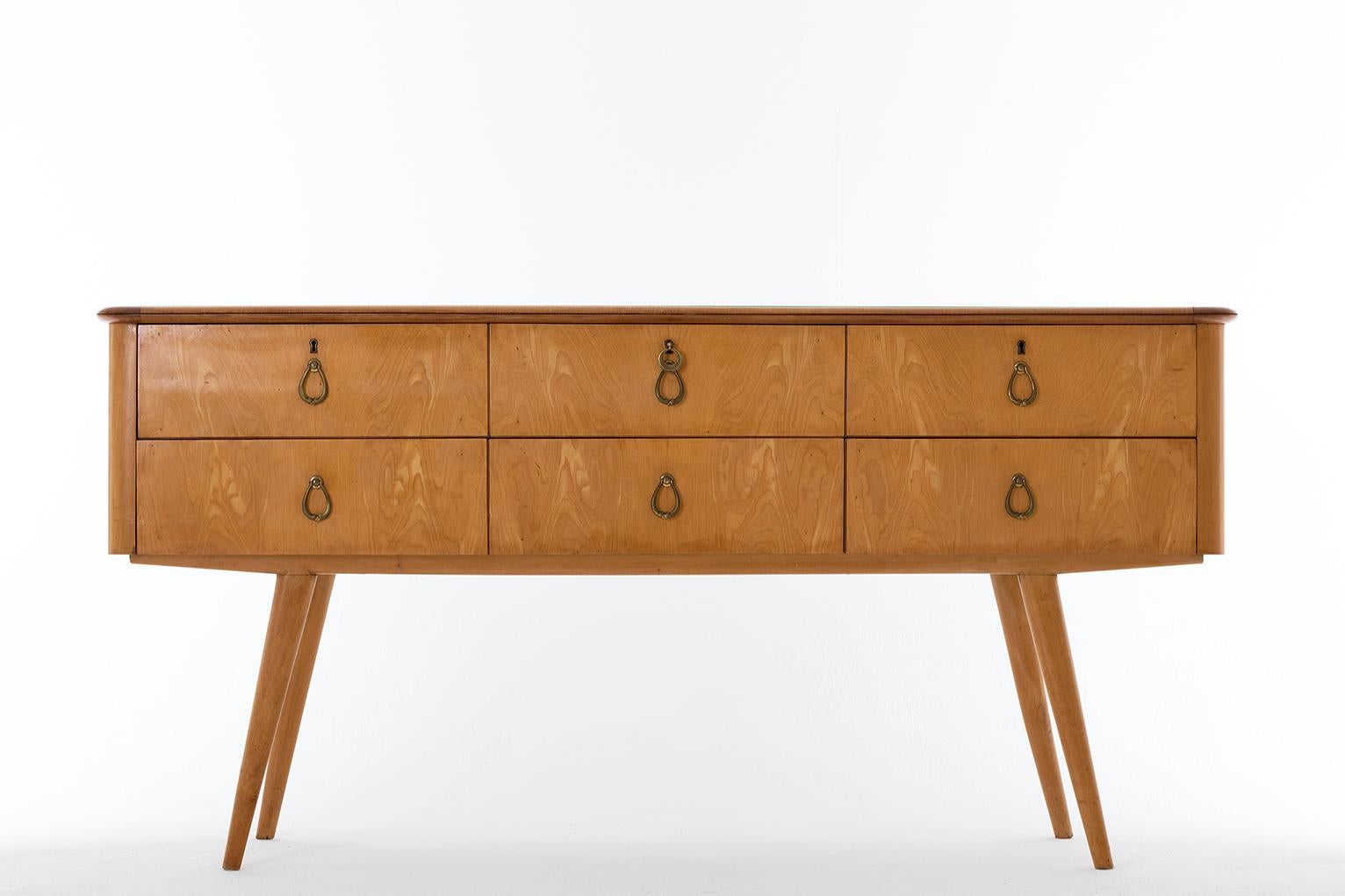 A large sycamore chest of drawers, the glass shaped top above six drawers with brass handles, with its original key, on four tapered and angled long legs,
Italy, late 1940s-early 1950s.