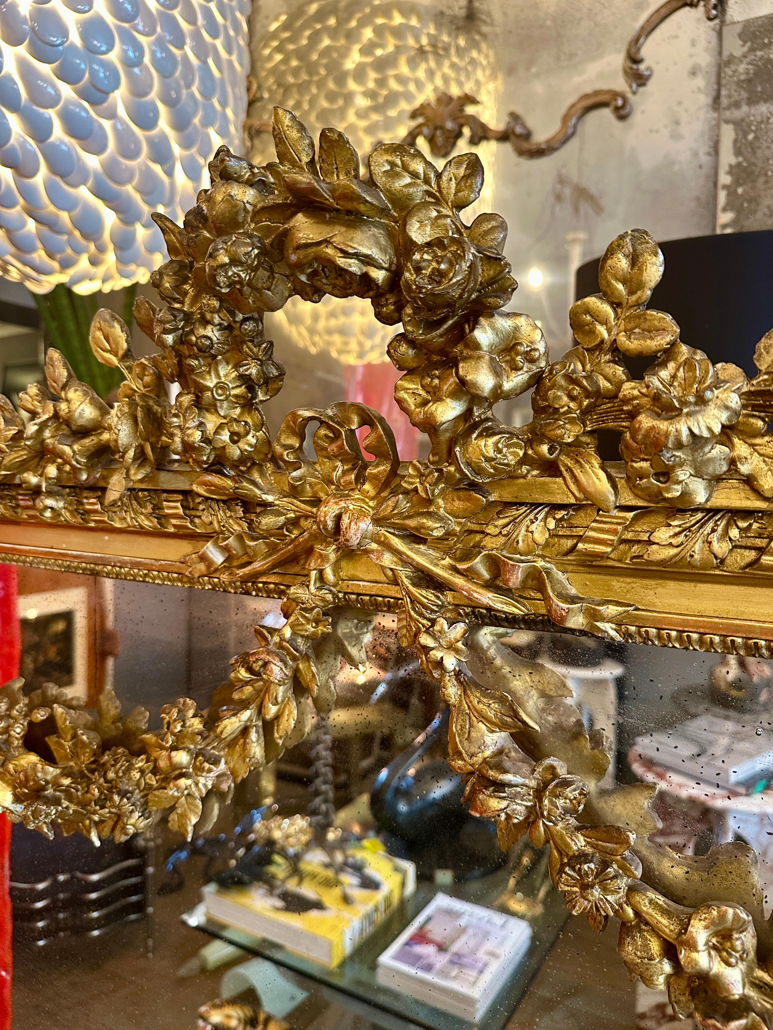 A sizeable ornate French Louis XVI style overmantel mirror. The top crested by a large floral foliate arch above a ribbon tied bow and quiver again with floral foliate swags to shoulders held by rosettes. The frame decorated with ribbon tied