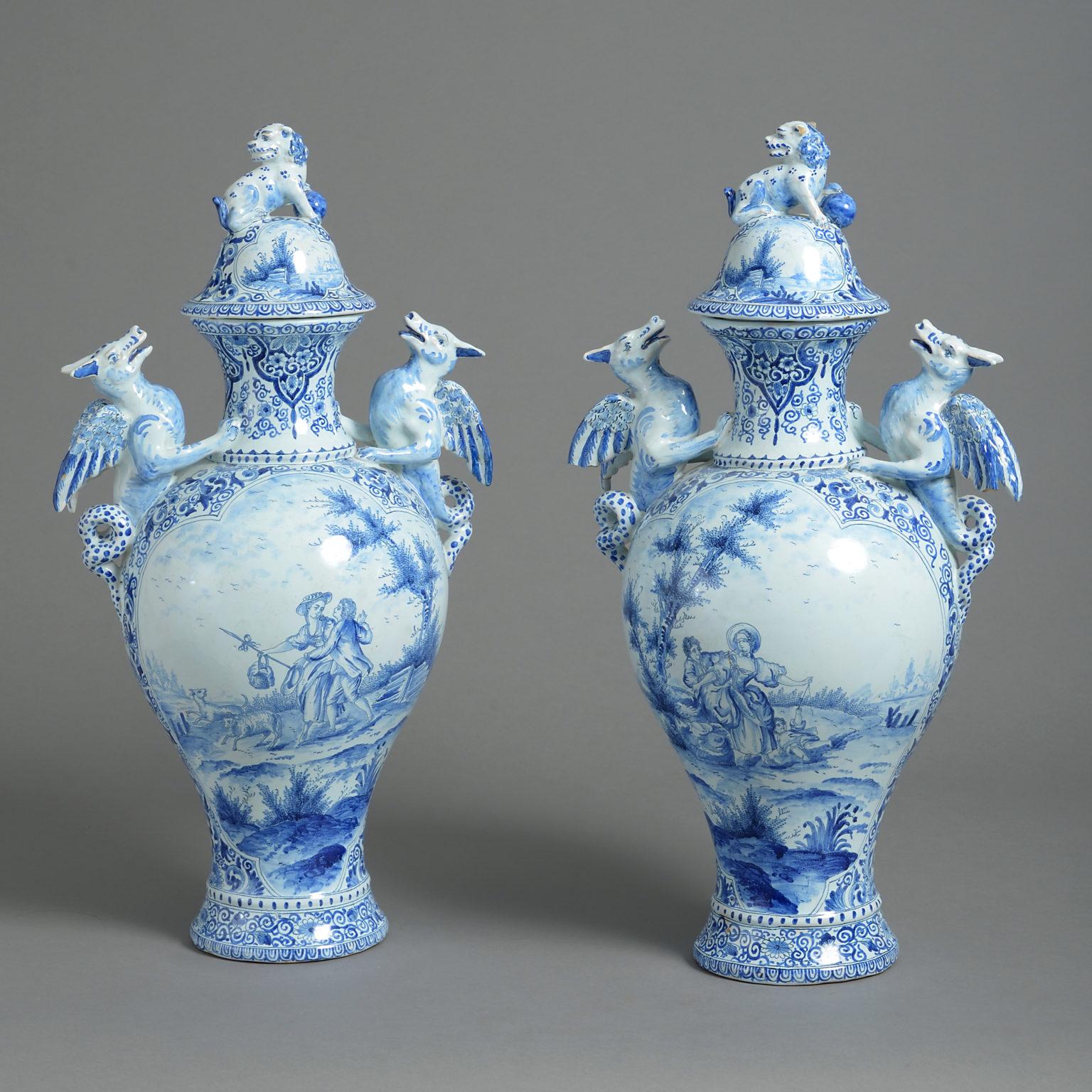A large early 20th century pair of blue and white glazed Delft pottery vases and covers, the lids surmounted with leopards and set upon bodies of baluster form, with dragon handles, all decorated with cartouches containing landscapes and figurative