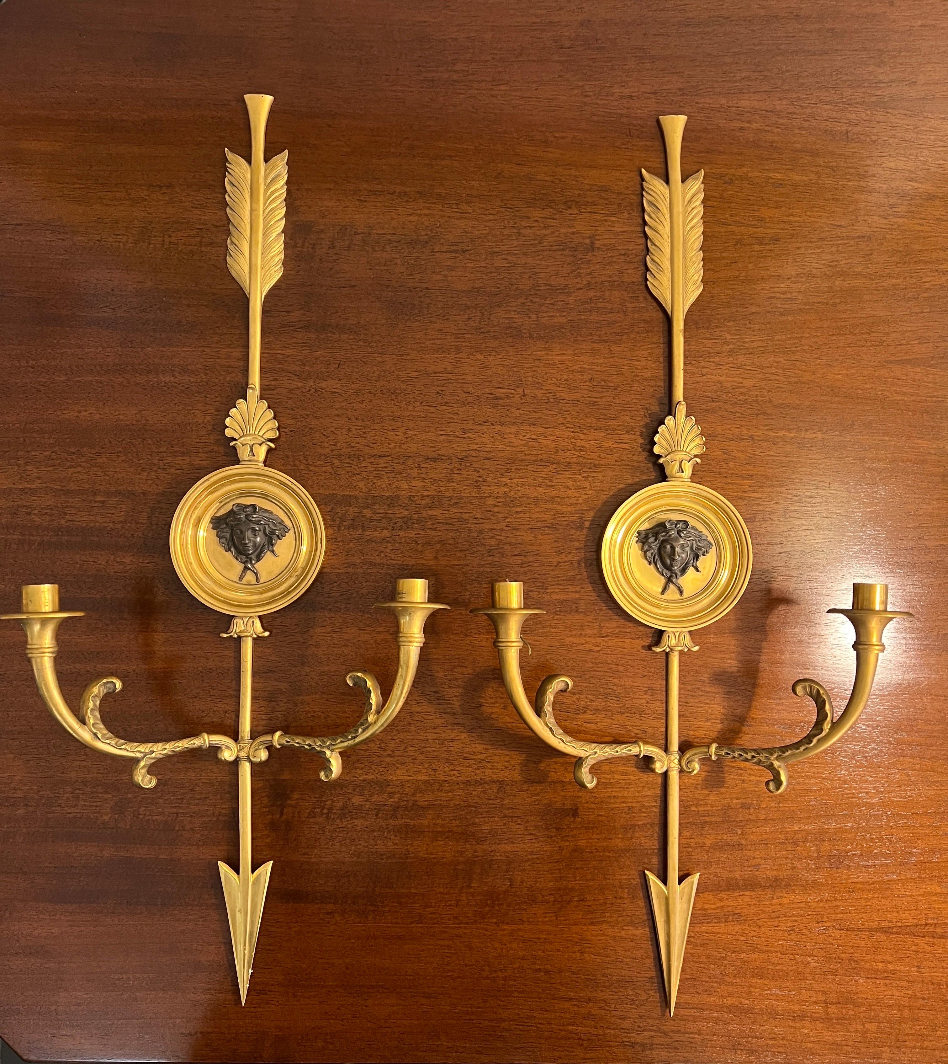 A magnificent large pair of early 20th century neoclassical ormolu arrow wall sconces. At the centre of each arrow is a decorative circular plate featuring beautifully detailed bronze relief of a woman’s face . 
The sconces are currently unwired