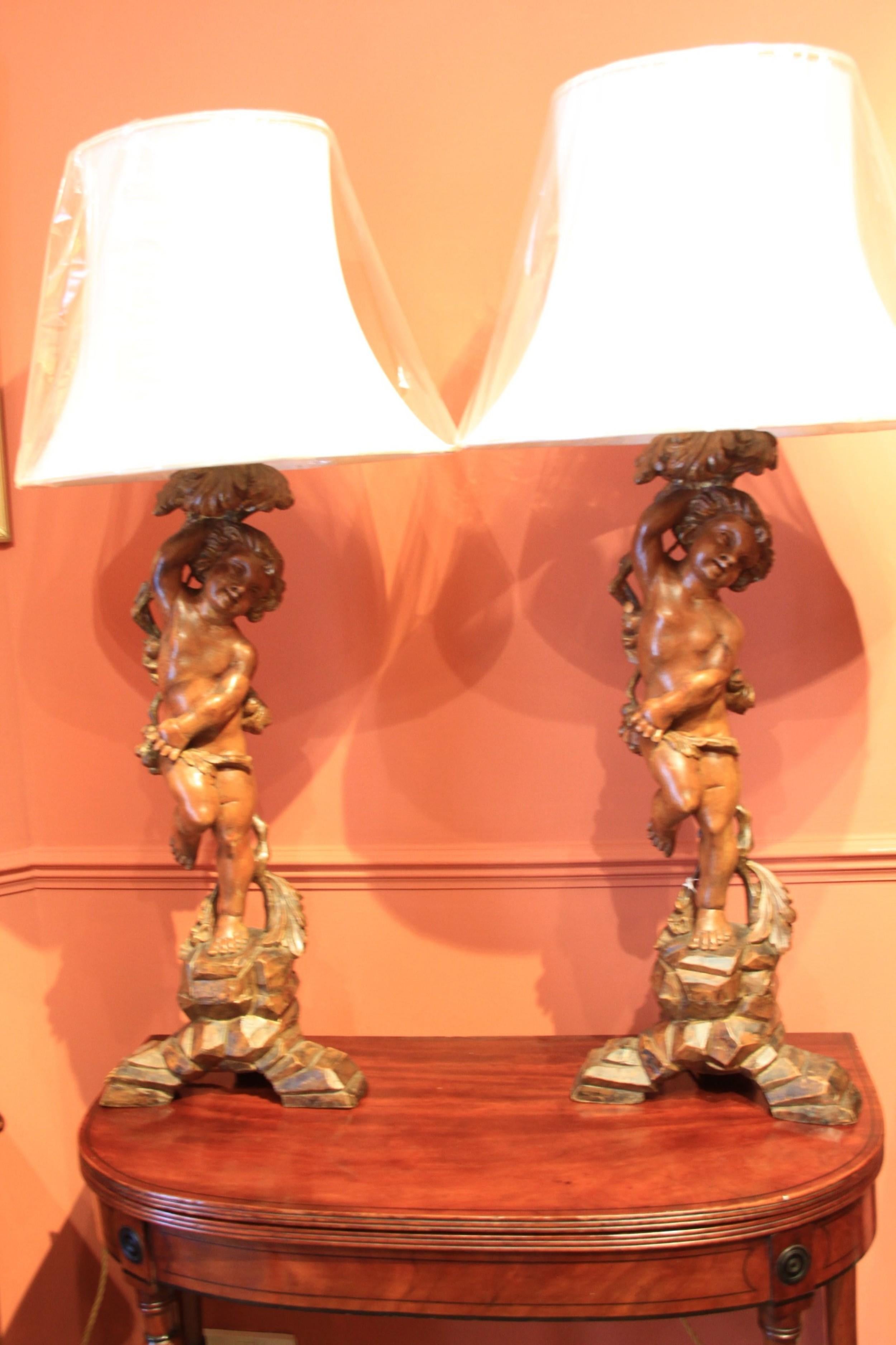 A highly decorative near pair of large impressive hand carved pine table lamps in the form of cherubs gathering grapes off a vine and standing on rock formed bases complete with a pair of cream silk oval shades.(new)

Measure: 107cm H including