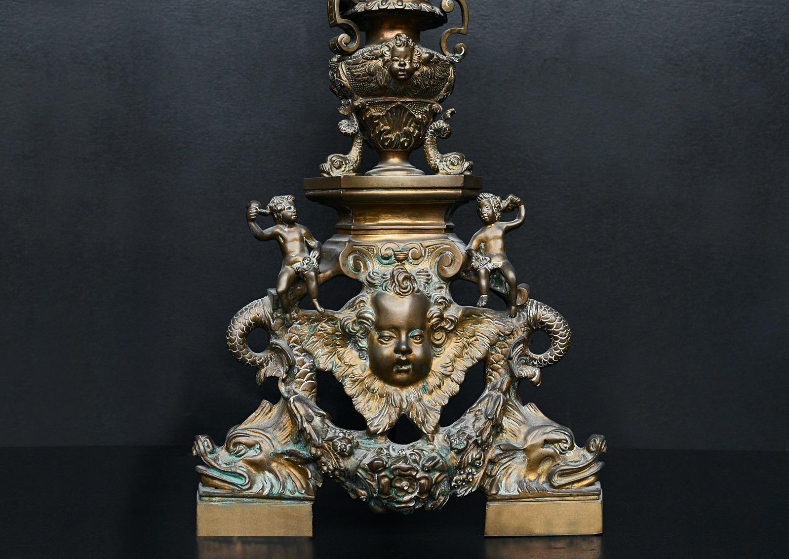 A large pair of ornate brass firedogs. The feet surmounted by dragons, foliage and putti, with urns to central shaft. Classical figures to top. English, 19th century. Currently with aged brass/bronze patina - could be polished to brass if