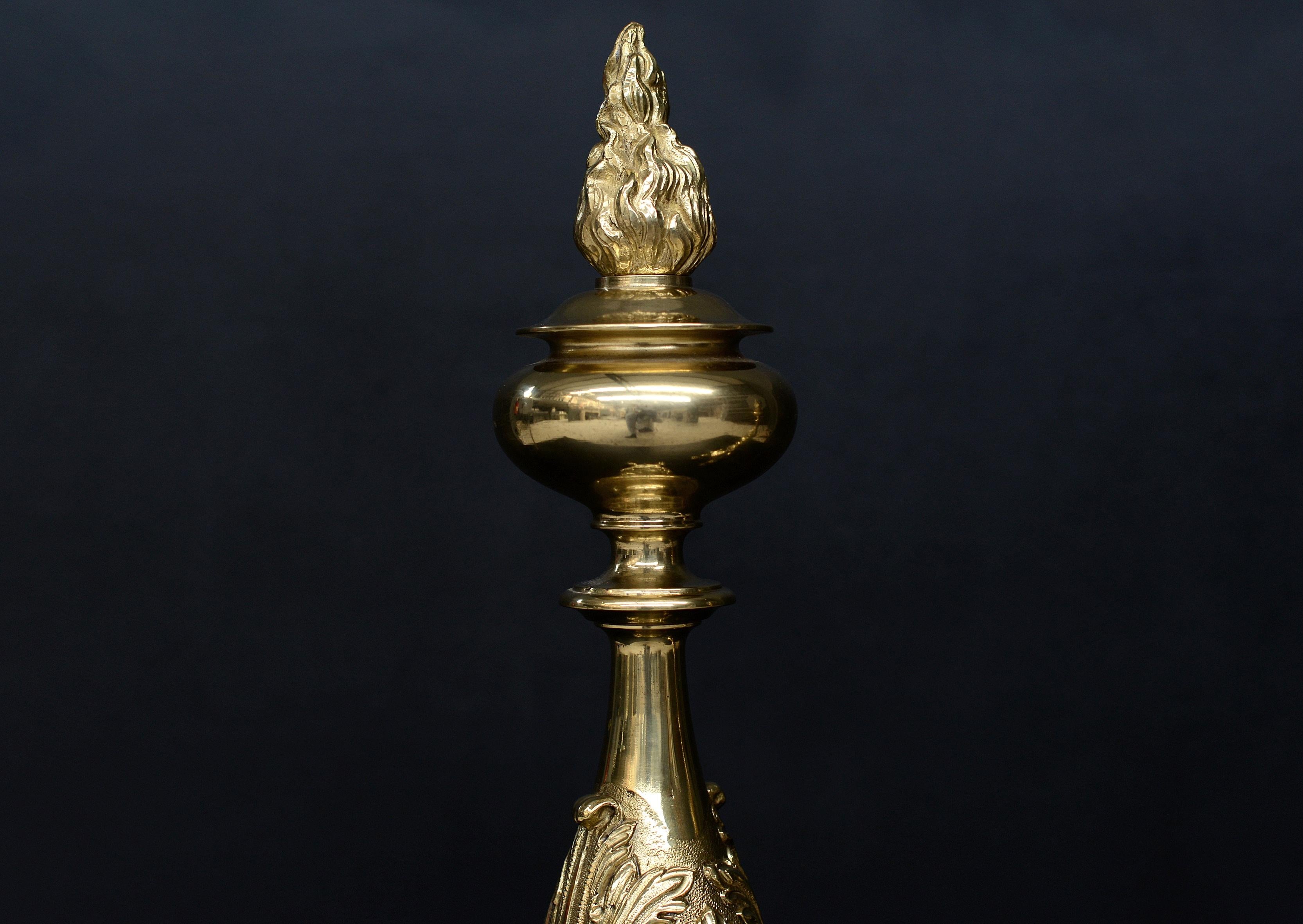 A large pair of brass firedogs. The bases of four clawed feet, the uprights with swags of fruit and stiff acanthus leaf detailing, surmounted by flamed finial tops. English, circa 1900.

Height:	865 mm      	34