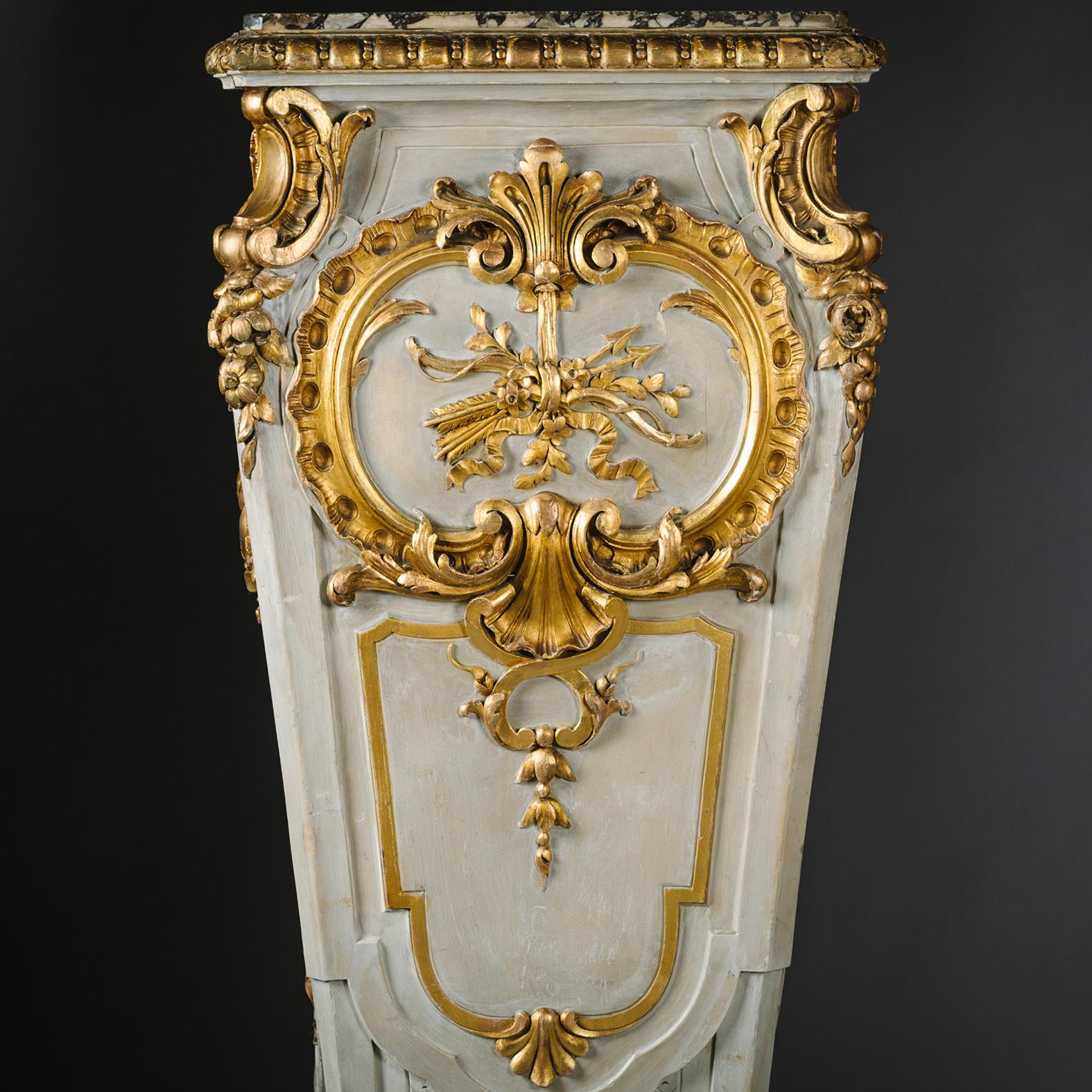 A Large Pair of Parcel-Gilt and Grey Painted Pedestals.

Each pedestal is of square tapering section inset with an Escalette Alpha rose marble top and carved overall with scrollwork, centred by an acanthus cartouche with ribbon tied trophy, above