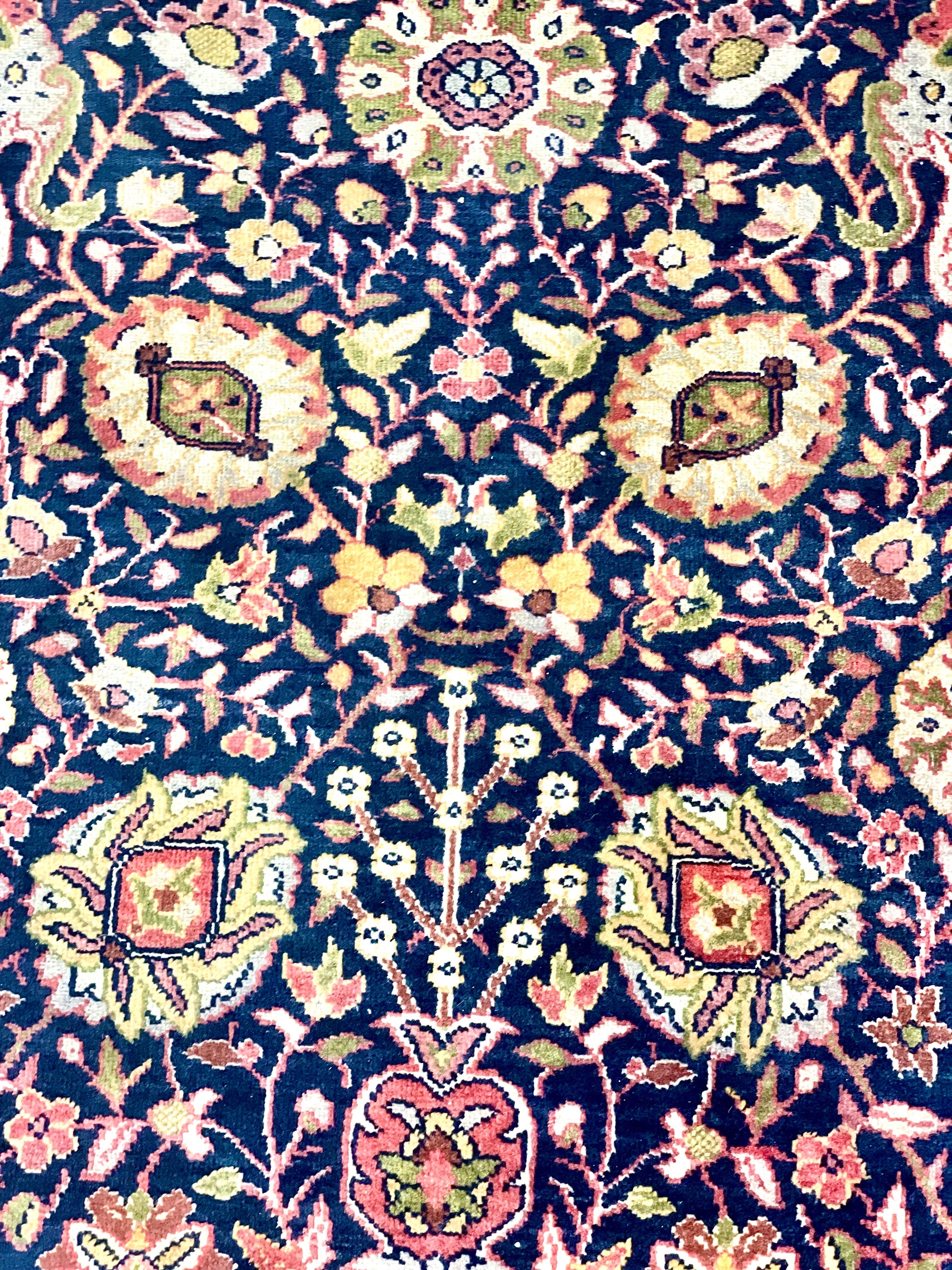 Un vibrant tapis persan, tissé à la main en pure laine. Ce très grand tapis merveilleusement décoré présente un fond bleu nuit orné de fleurs et de feuillages stylisés entrelacés. La bordure rouge contraste parfaitement avec le centre plus foncé et