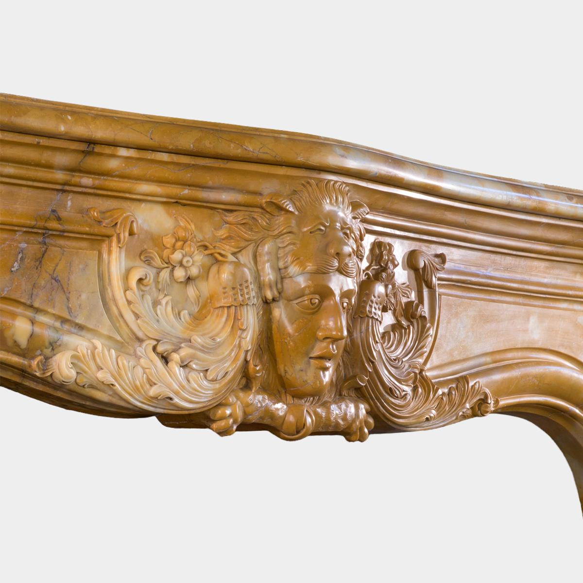 A large chimneypiece executed in yellow Siena marble. The central cartouche carving Hercules  wearing the Nemean lionskin, flanked on either side by carved foliate scrollwork, which is reflected on the corners of the panelled frieze and again on the