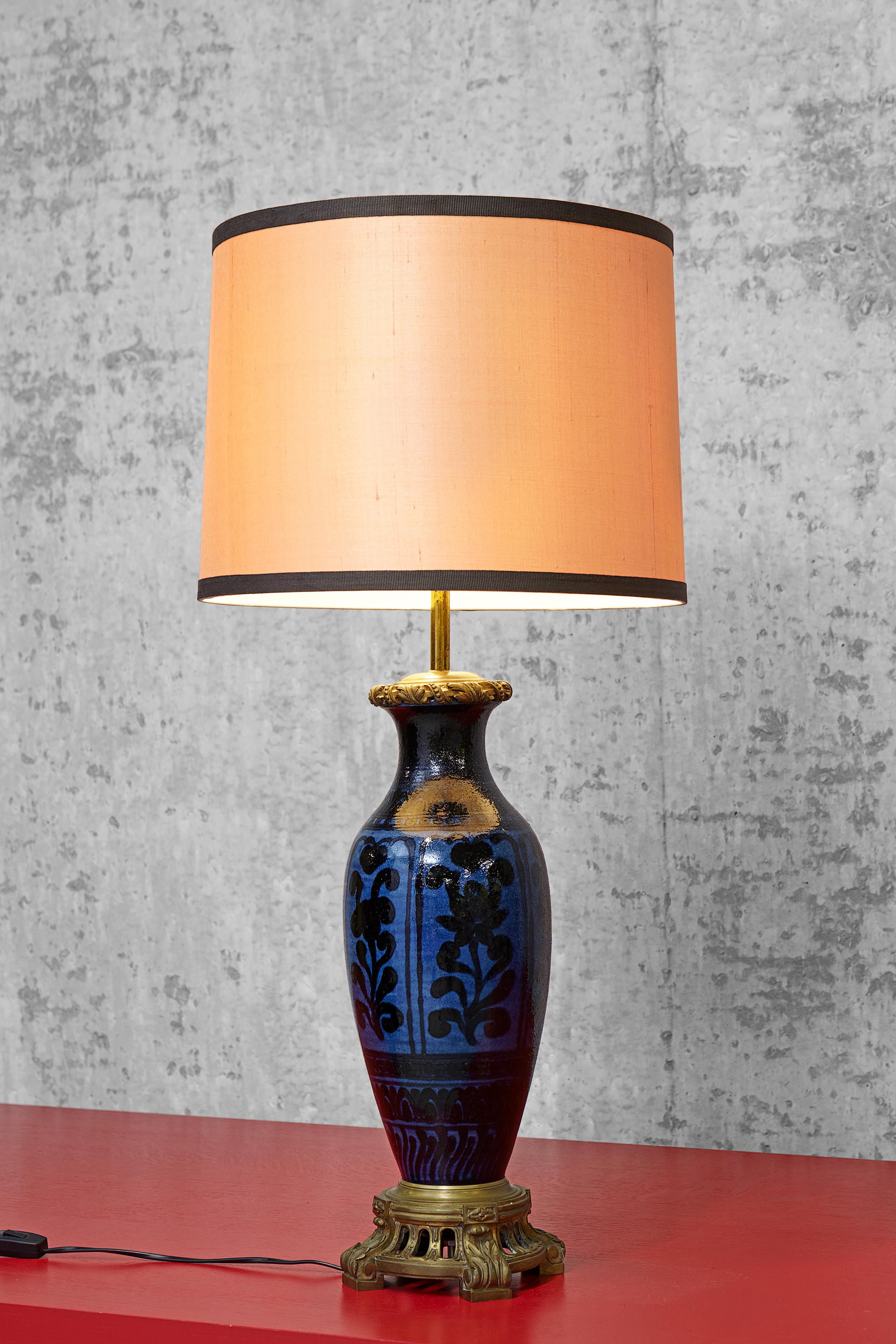 A large table lamp in the form of a vase. The dark blue ceramic vase is decorated with a black leave and flower pattern in Persian style. The vase is attached to a round moulded brass stand on four legs, decorated with acanthus leaves in the style