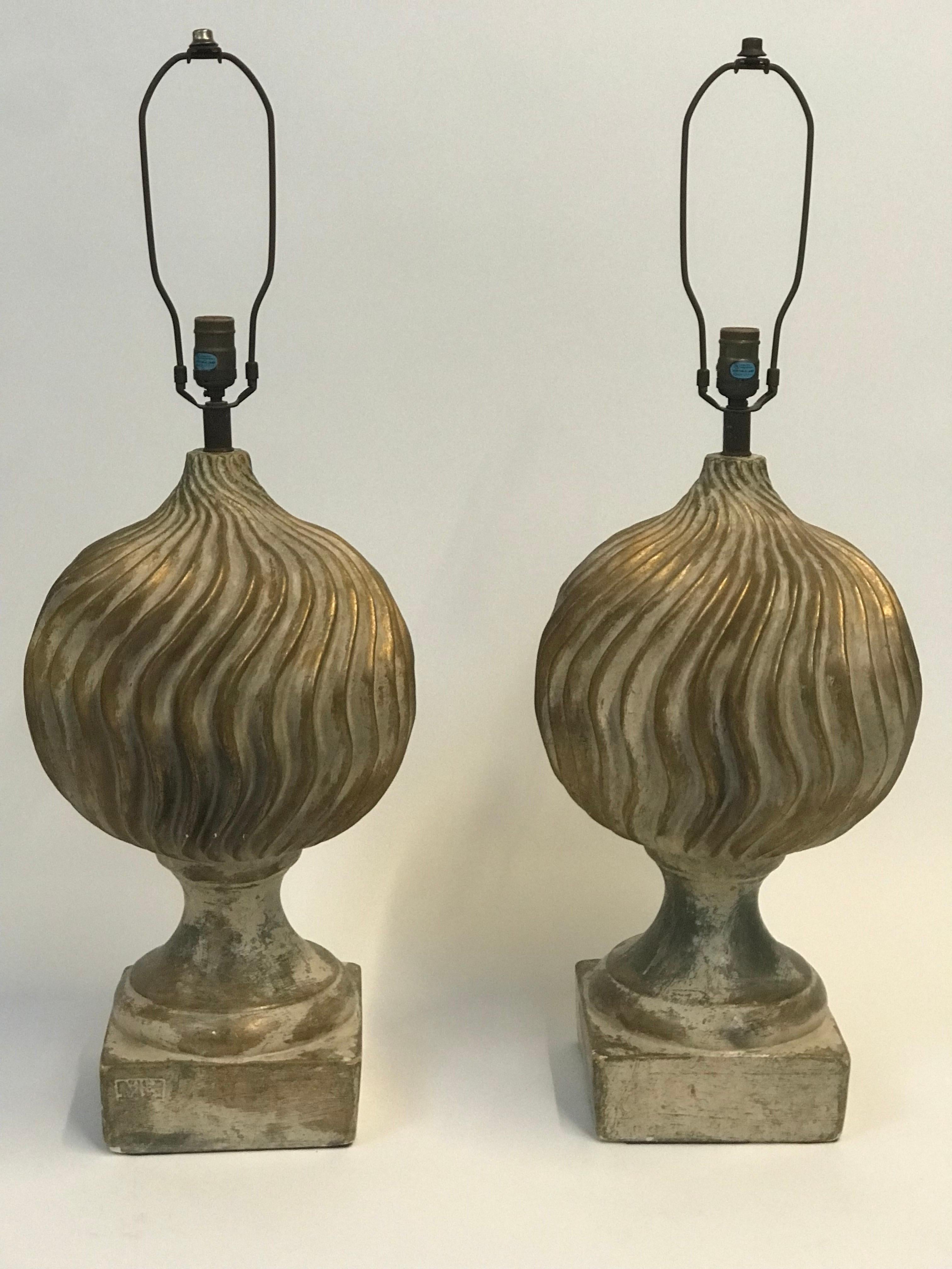 A large format pair of plaster table lamps in a 1950’s stylised neoclassical form consisting of a complex wave ribbed globe rising from a traditional form plinth like base and finished in a rubbed distressed gilt semi metallic glaze with inclusions