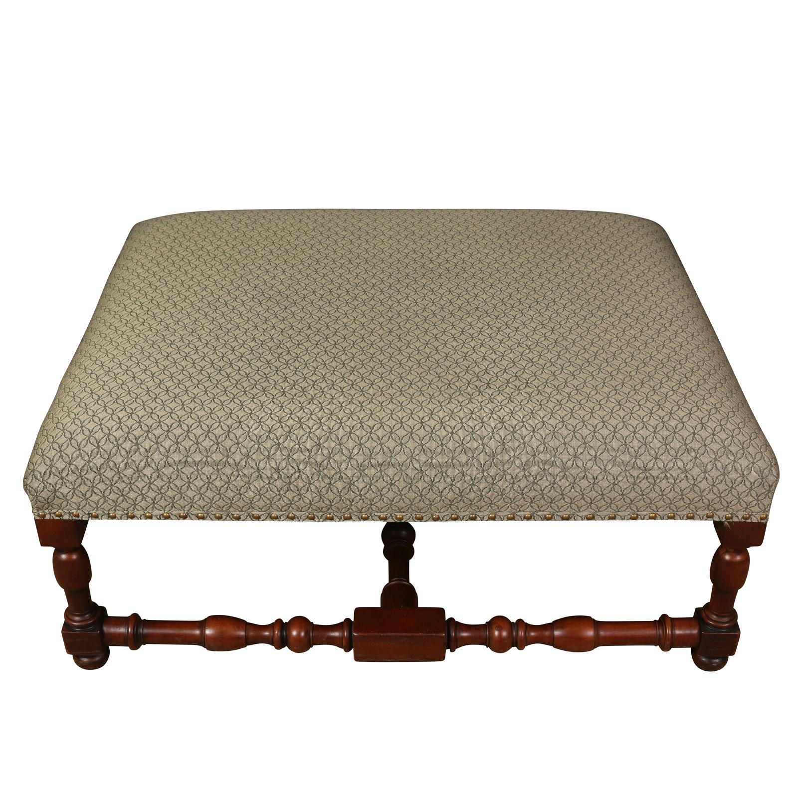 A nice looking square ottoman with an upholstered  top and a turned leg base with a cross stretcher.  Upholstered in a neutral fabric and finished with a tape and nail heads, this handsome piece can also serve as a coffee table for casual