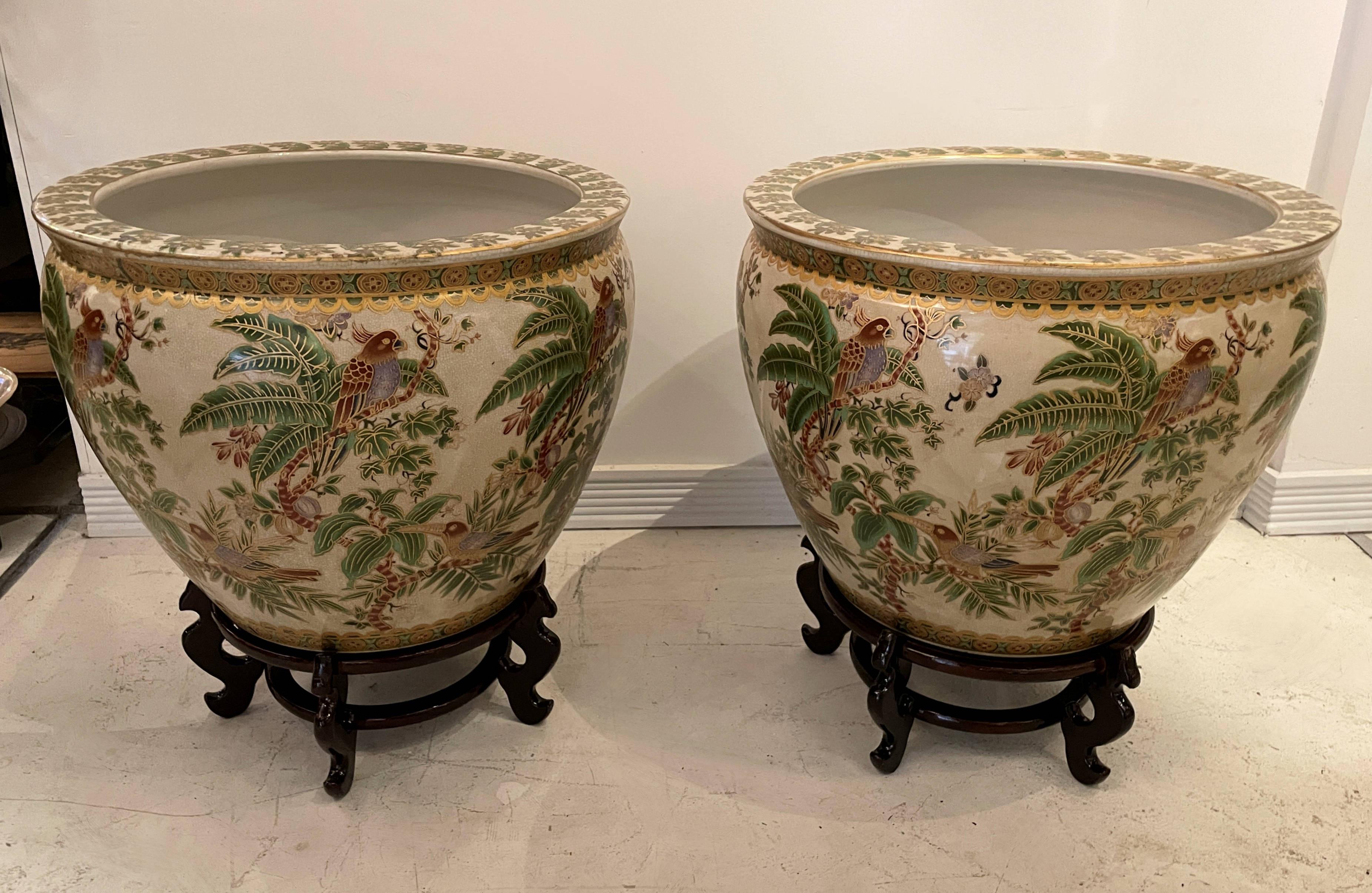 Vivid tropical scenes are hand painted on a pair of Chinese porcelain fishbowl planters with matching wood stands. The interior is delightfully painted with coy fish. The pair are in very good condition.