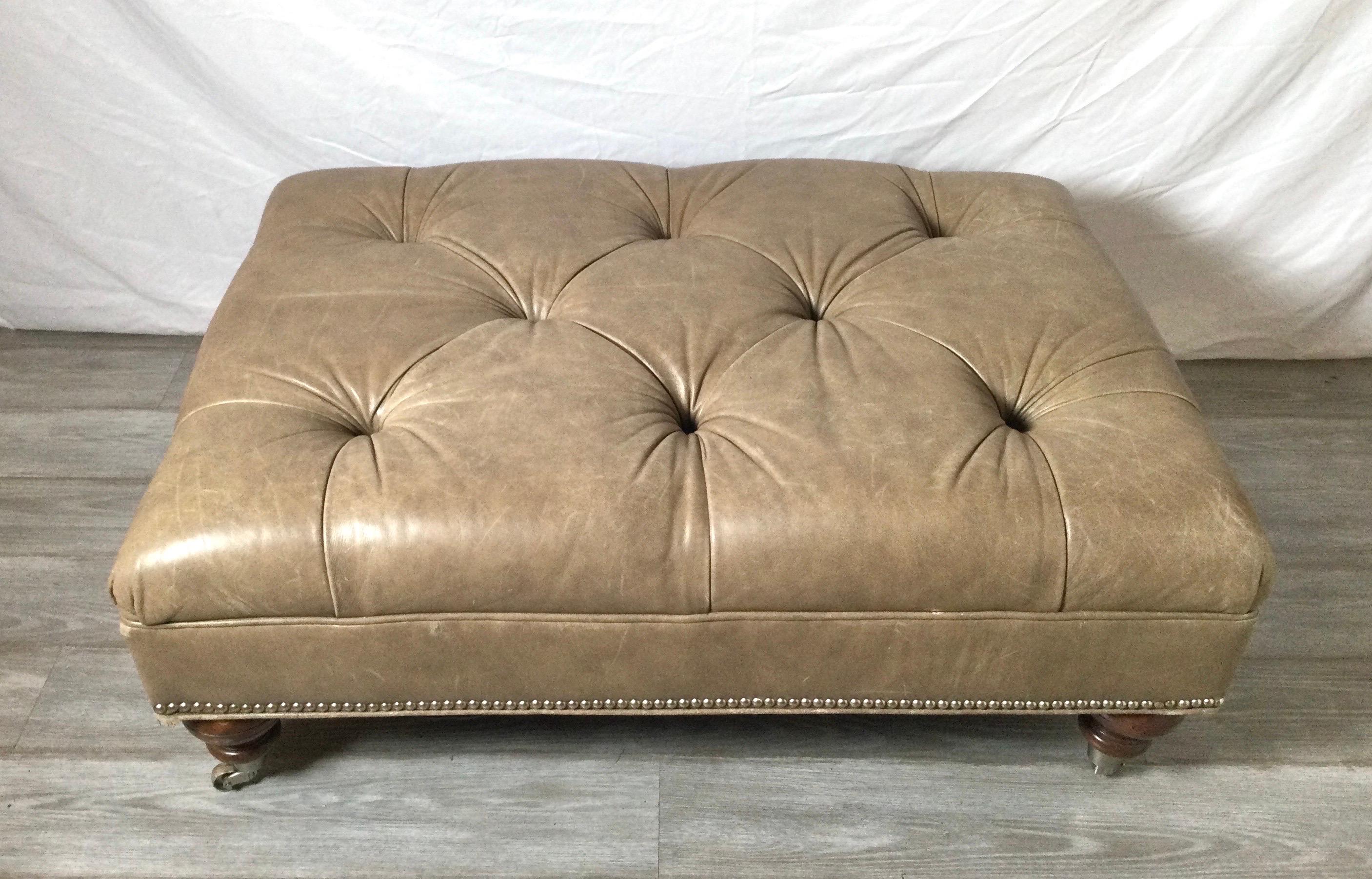 A large Taupe leather tufted ottoman on turned walnut legs with brass castors. The genuine leather with brass nail-head trim all around. The large size is perfect as an alternative to a coffee table, easy to move when needed.