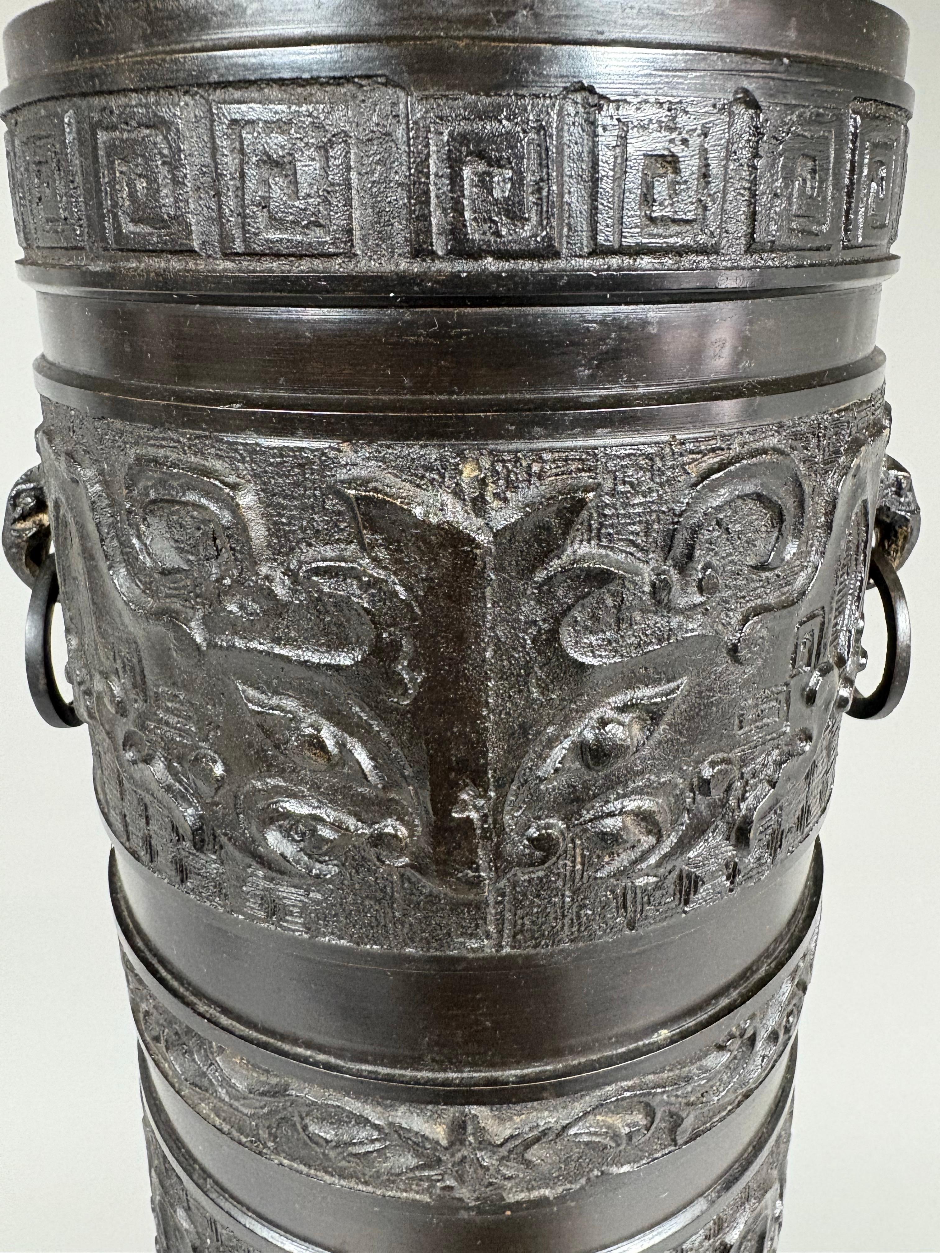 Lampe en laiton de la période Hollywood Regency d'une forme de colonne d'inspiration chinoise, attribuée au style de James Mont. Le corps est décoré de bandes horizontales archaïques, comprenant des masques de taotie stylisés, et est flanqué sur