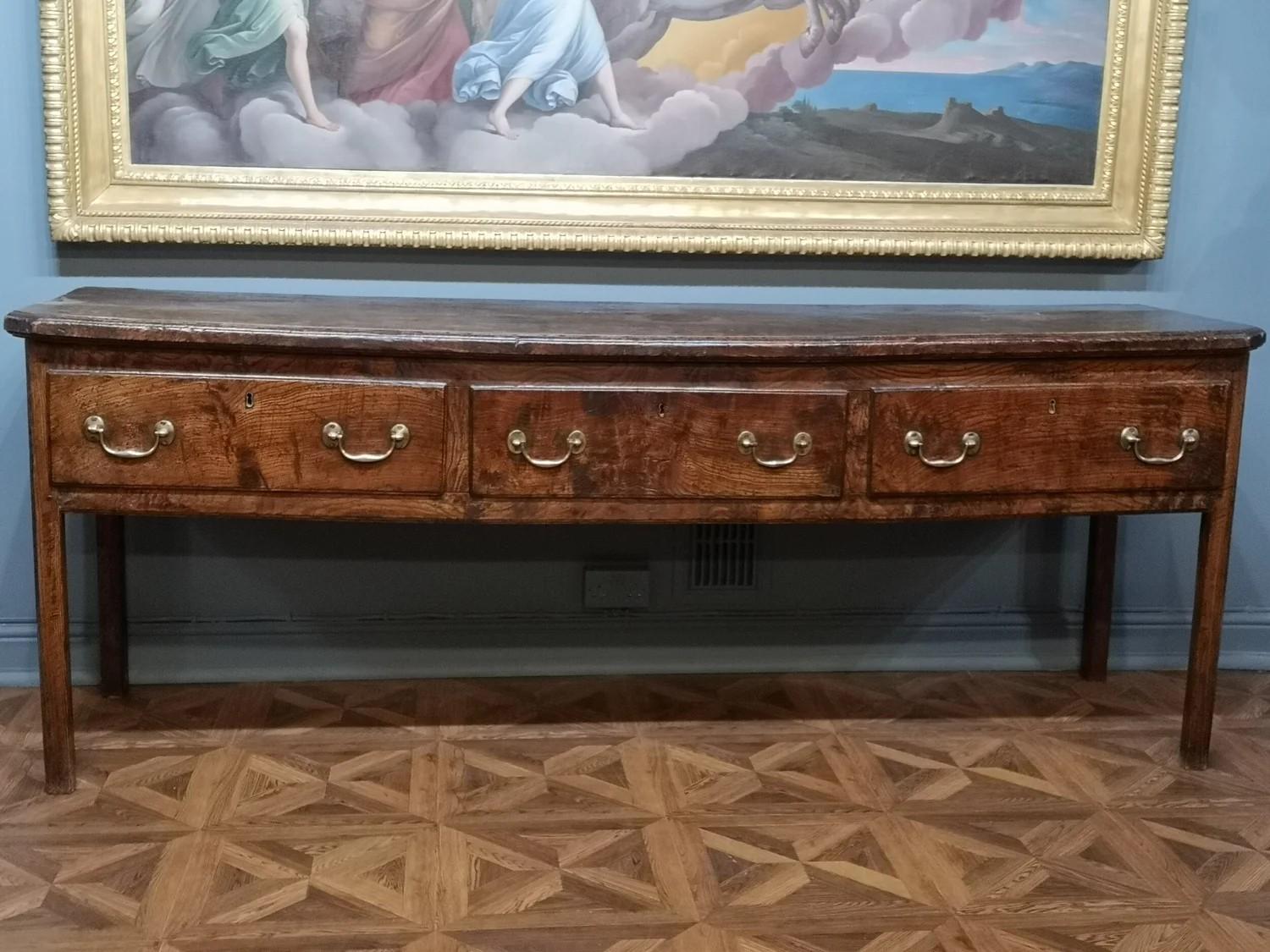 Comò basso in olmo della fine del XVII secolo di grandiose proporzioni. Di forma aperta con tre cassetti sopra le gambe quadrate, le gambe anteriori con una semplice nervatura,  la parte superiore presenta angoli anteriori inclinati e  Bordi