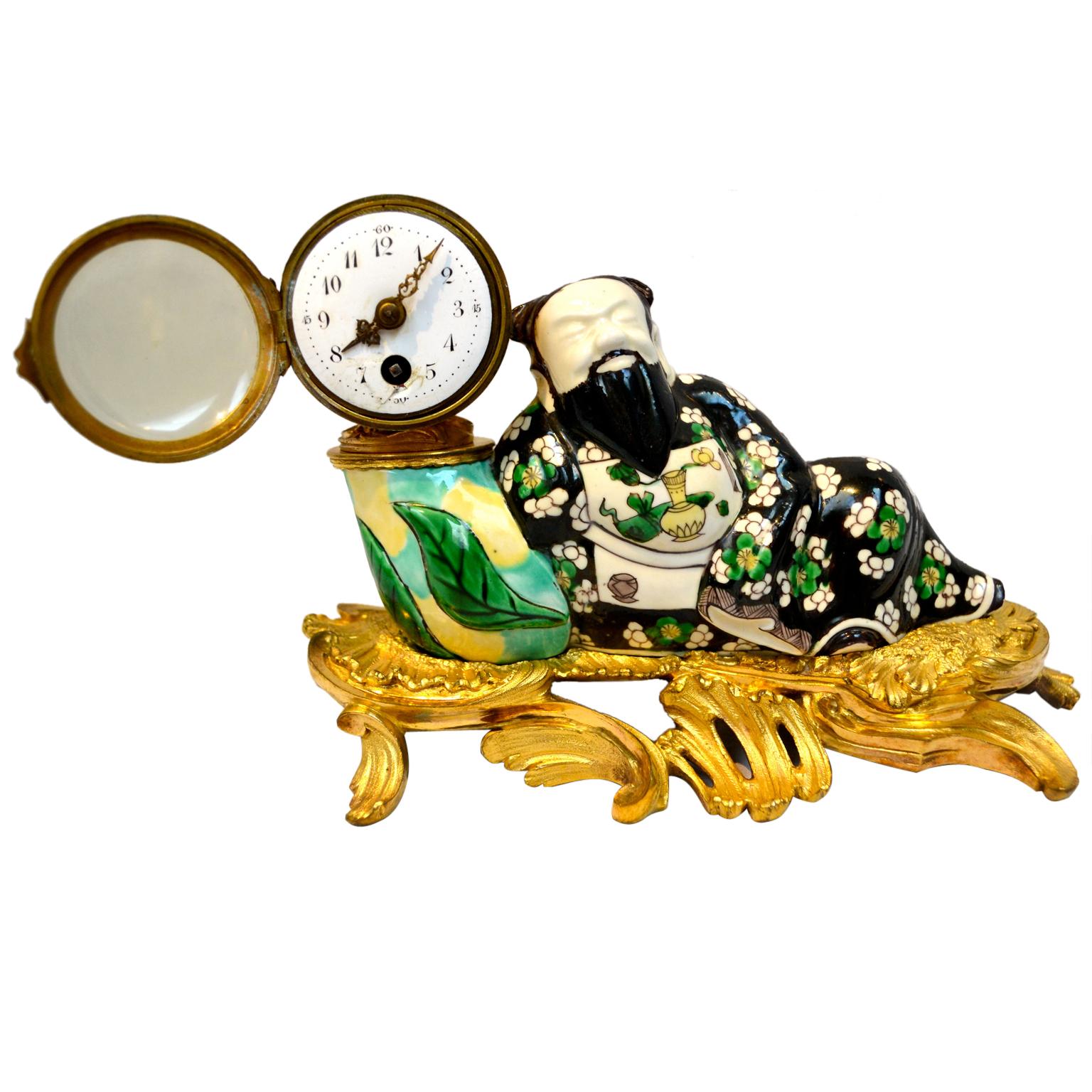 A finely modelled and painted Louis XV-style porcelain desk clock with a figure of a recumbent Chinese man (wise man). He leans against what appears to be a tobacco drum, of which a Swiss made clock or watch movement is mounted. The Chinese