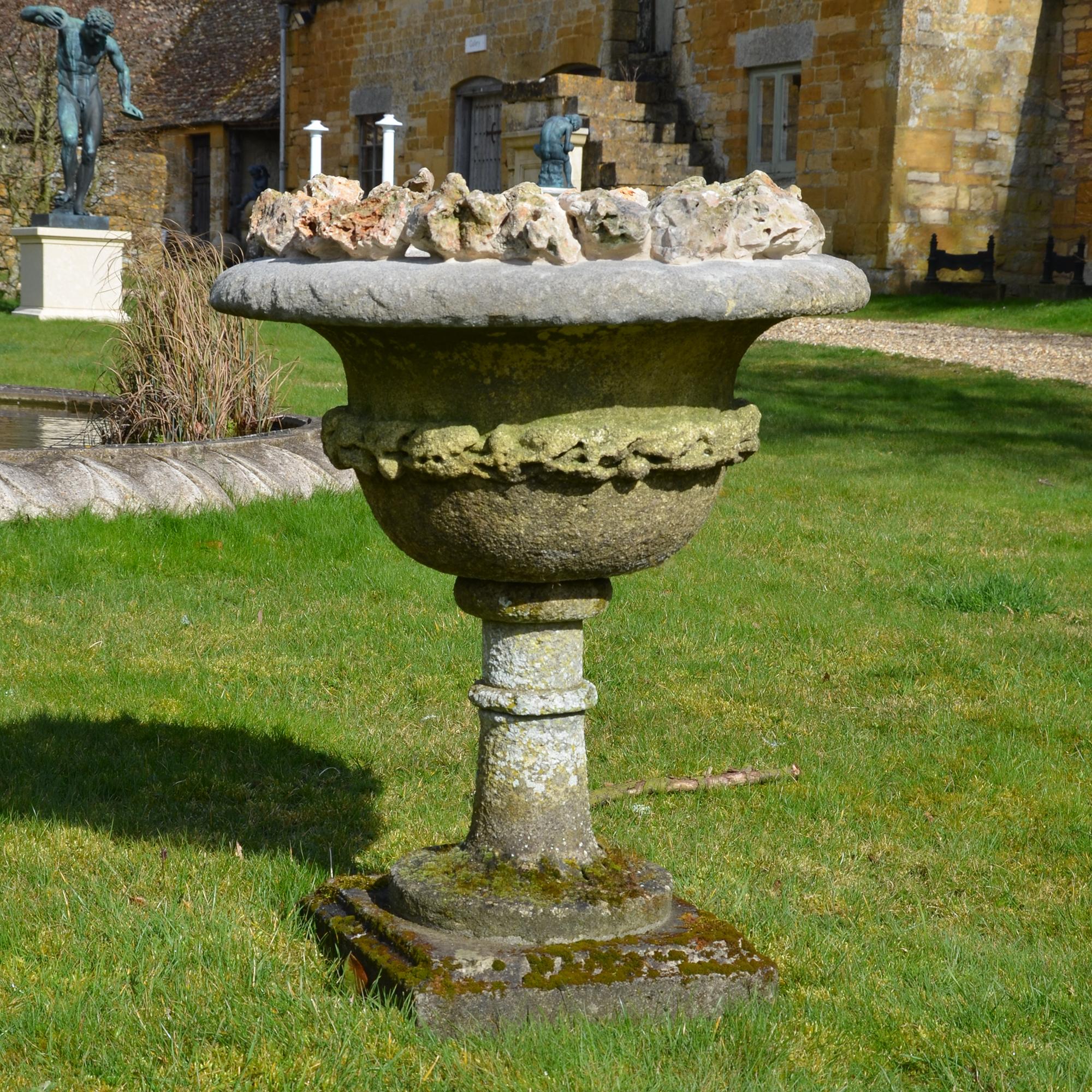 A late 19th century carved stone urn of campana form having a foliate decorative band and later additional grotto stone decoration to the top rim, raised on an extended socle base.
