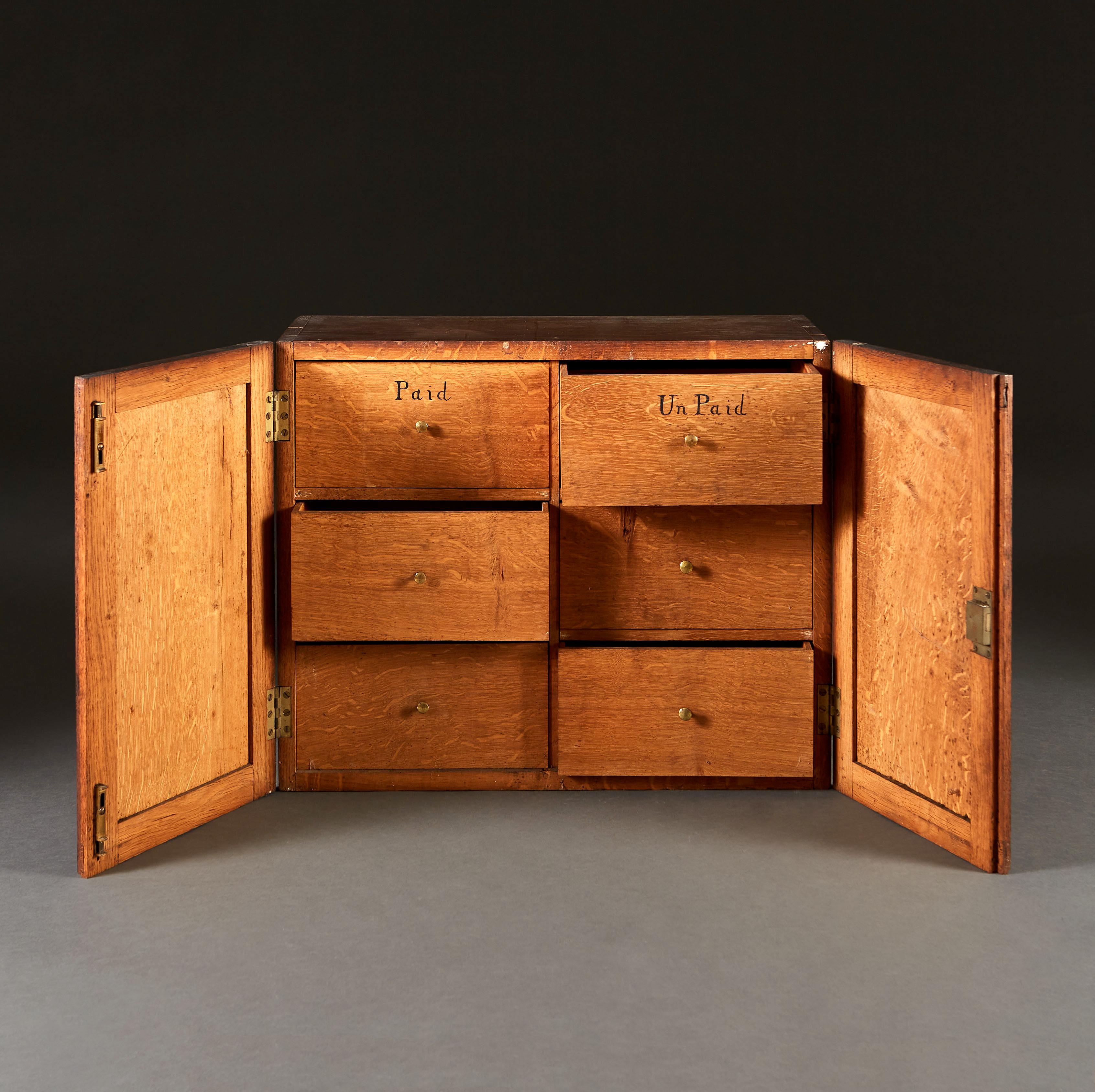 A late nineteenth century table top filing cabinet, probably from an estate office, with two cupboard doors opening to six drawers, the cupboard doors marked with the initials F.C.M. and the drawers marked for paid and unpaid invoices.