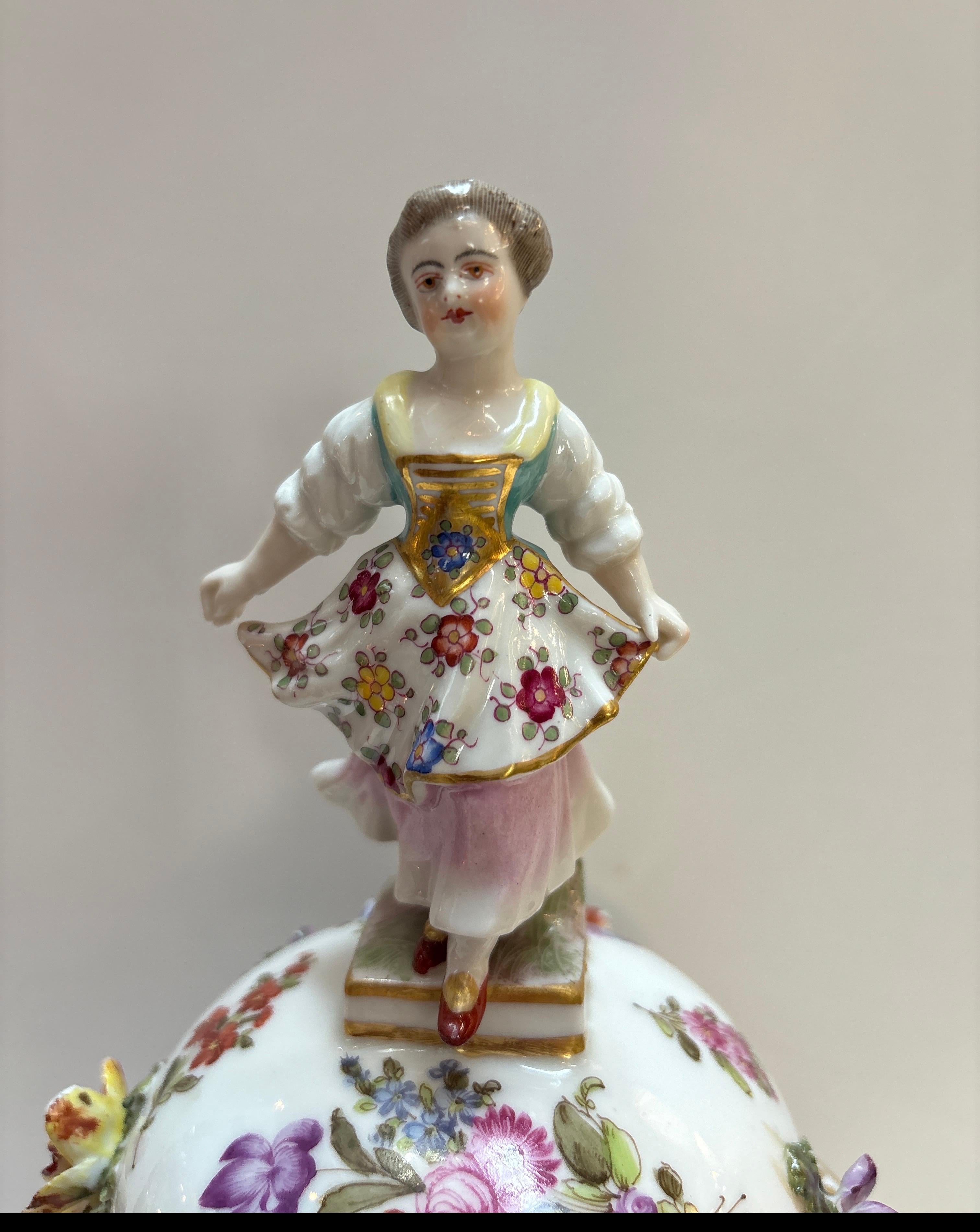 An elaborate hand painted porcelain, dressed and earn with lid. The lid with a figure of a young girl with hand painted floral decoration.  The main portion of the urn with three dimensional garlands of flowers all around.  15.25 inches tall, 8.5