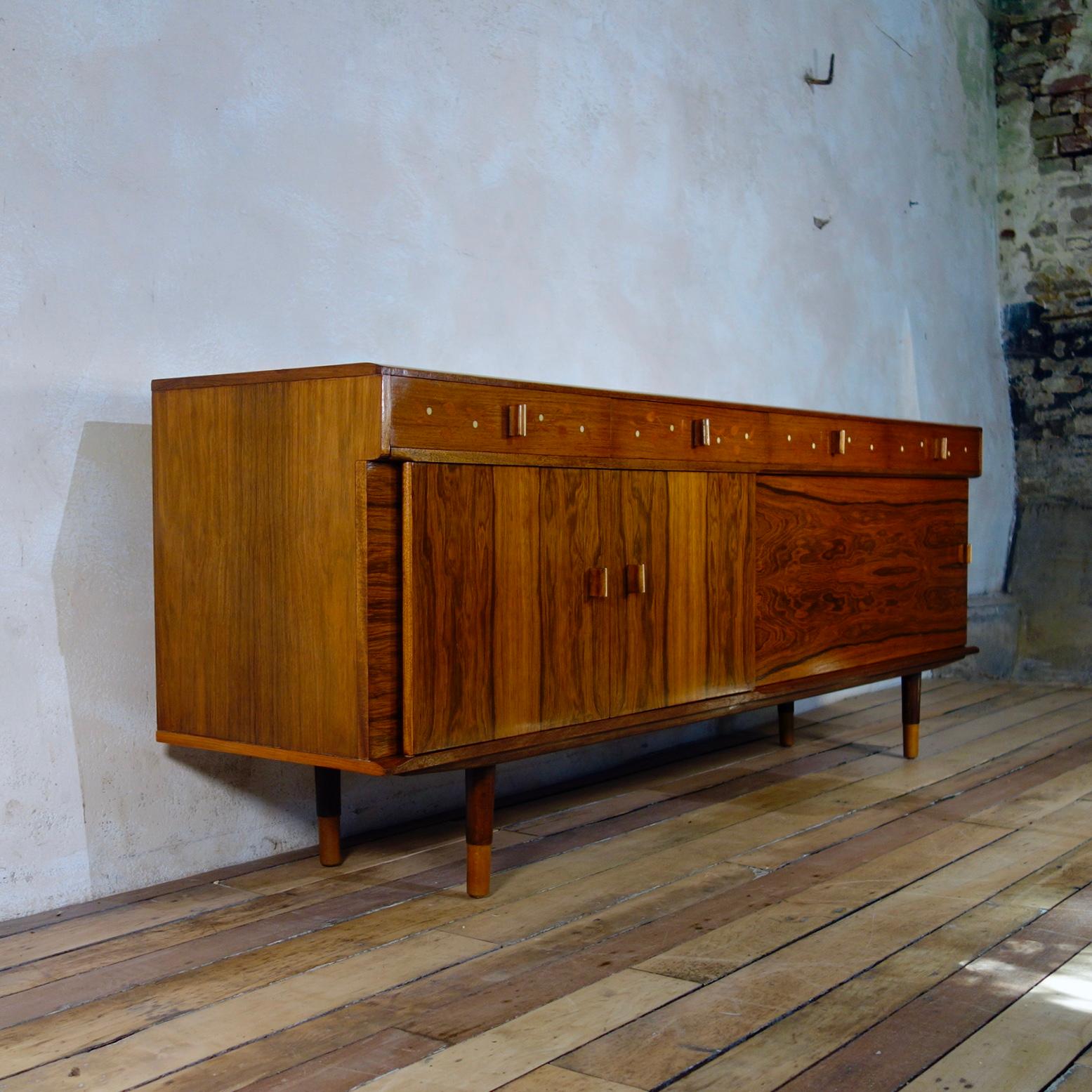 A Brazilian rosewood and inlaid sideboard/credenza.
Featuring four frieze drawers all of which have been subtly inlaid with repetitive mistletoe decoration. The far left-hand side drawer is lined with green felt and sectioned into four equal sized
