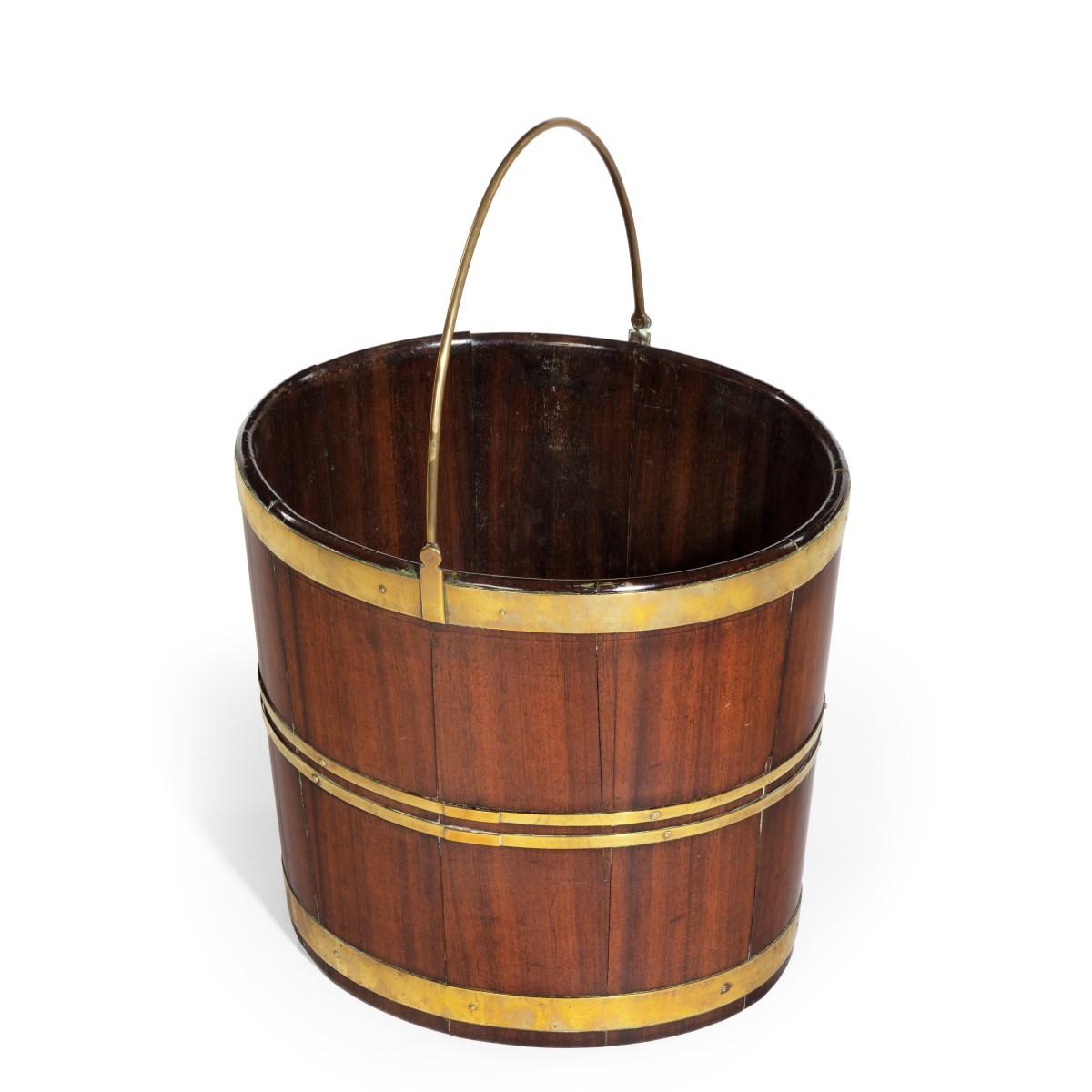 A late George III brass bound bucket, of oval shape with the mahogany timbers held by brass bands and with a hinged brass handle. English, circa 1810.
     