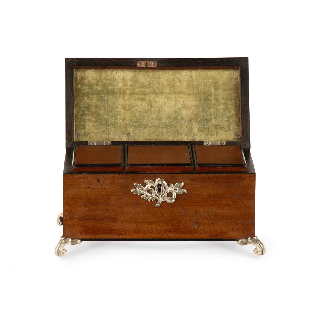 A late George III mahogany tea caddy, of rectangular form the hinged top opening to reveal three compartments with sliding lids, with silver mounts including a foliate scroll handle, an asymmetrical key escutcheon and four scroll feet,  and ebony